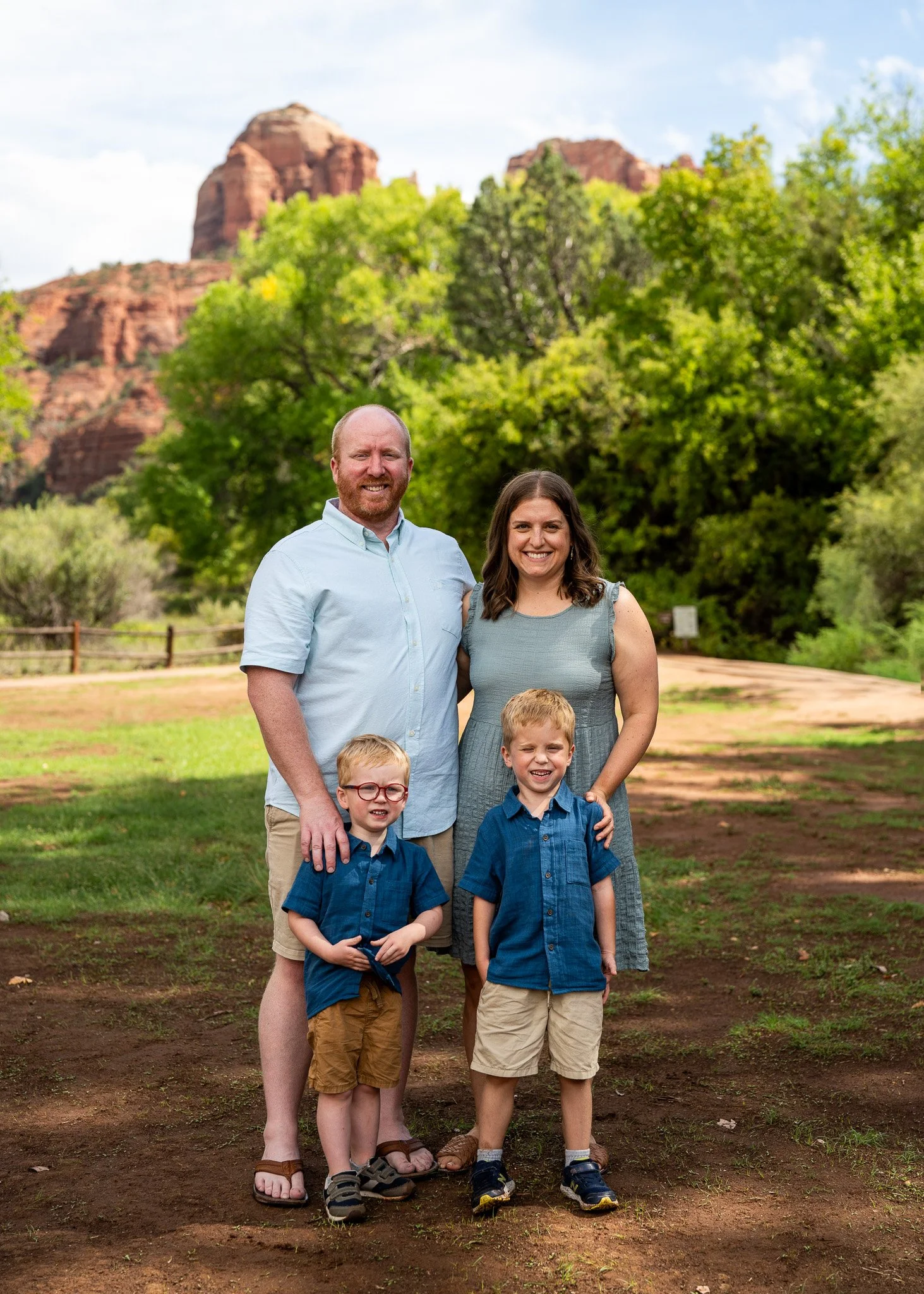 A family of four standing outdoors in front of green trees and red rock formations. The father is bald with a beard, wearing a light blue shirt and shorts. The mother has shoulder-length brown hair, wearing a gray dress. The two young boys are wearin