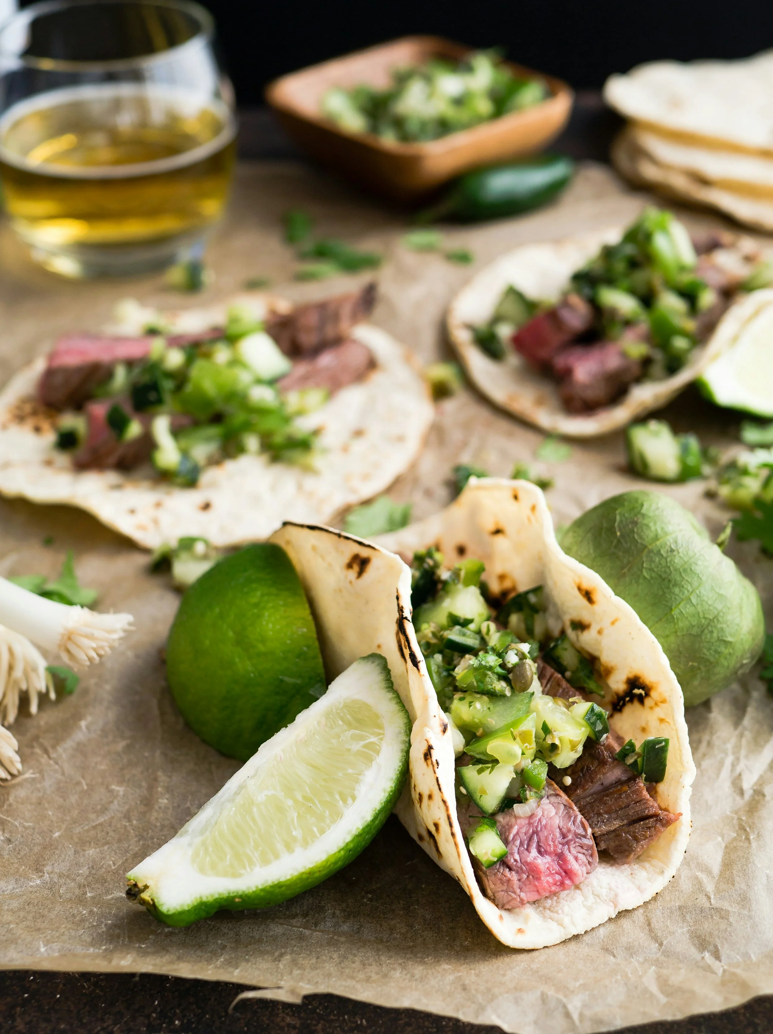Grilled steak tacos with lime slices and fresh cilantro on parchment paper, alongside a glass of beer.