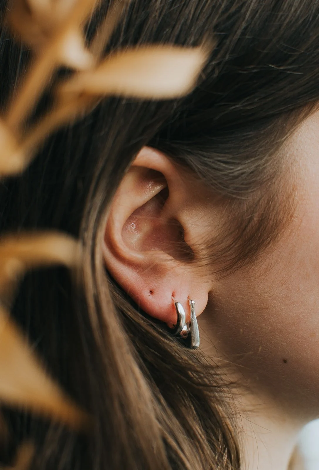 Close-up of a woman's right ear with a small hoop earring and a helix piercing, showing her brown hair and part of her face.