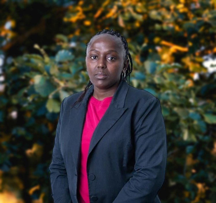 A woman with braided hair wearing a dark blazer over a pink shirt standing against a plain white background.