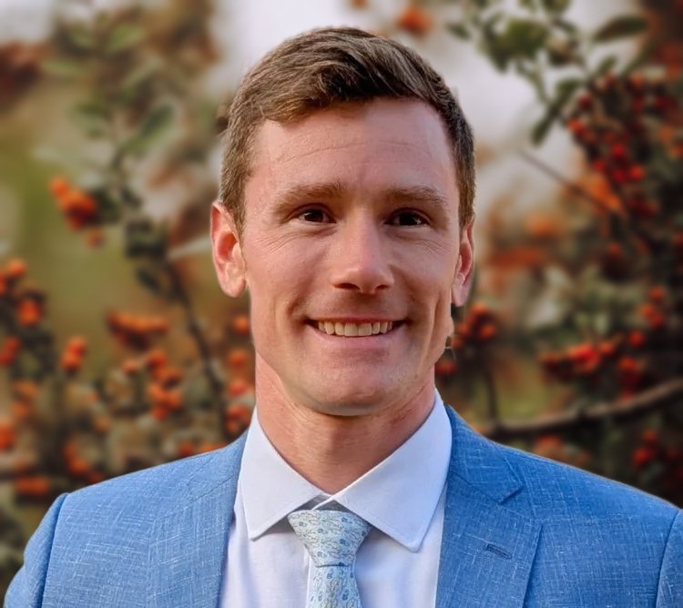 A young man in a light blue suit and white shirt smiling outdoors with a building and garden in the background.