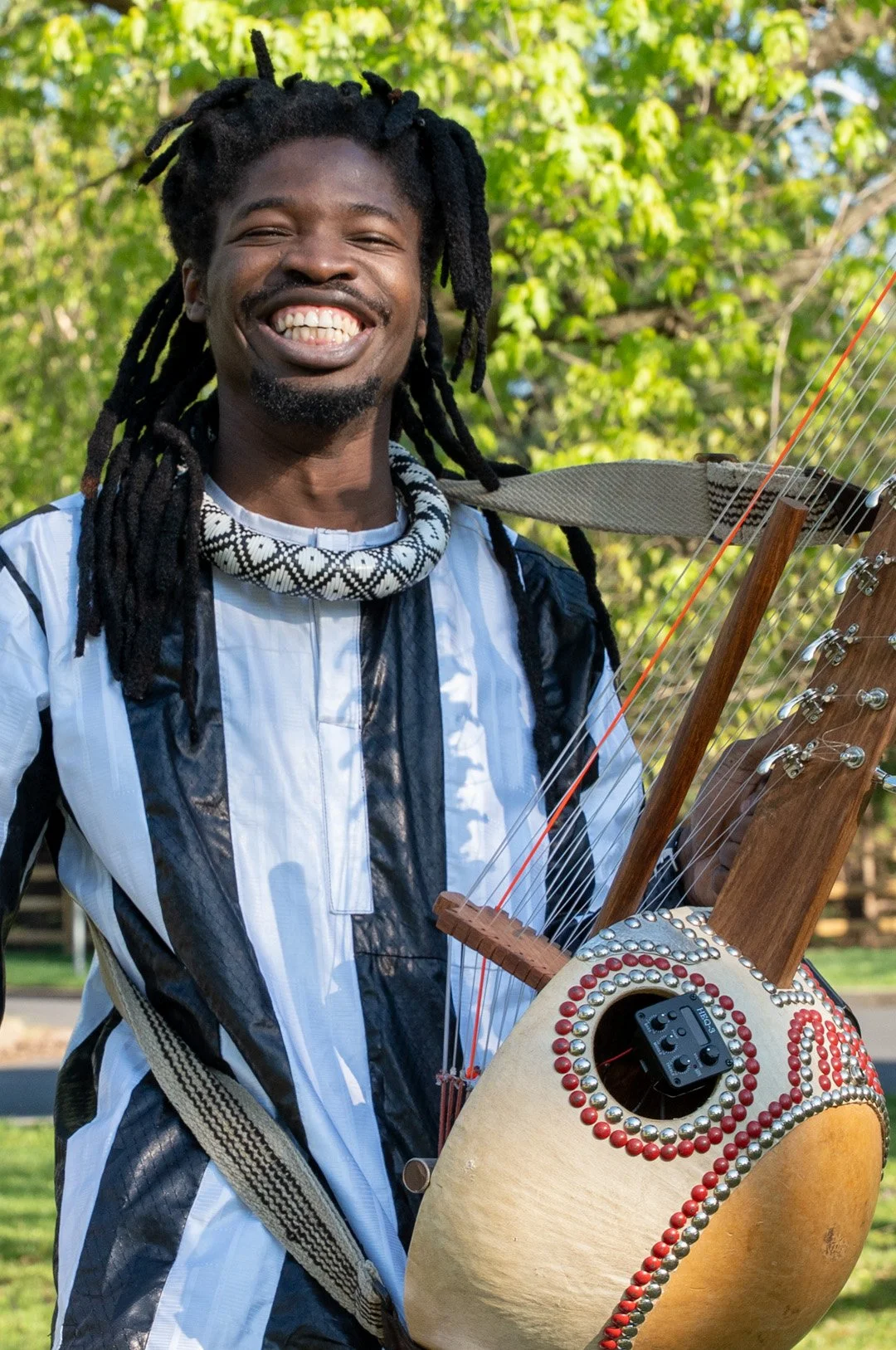 A young person with dark skin smiling widely, showing teeth, wearing a traditional Senegalese outfit, holding a kora against a background of trees.