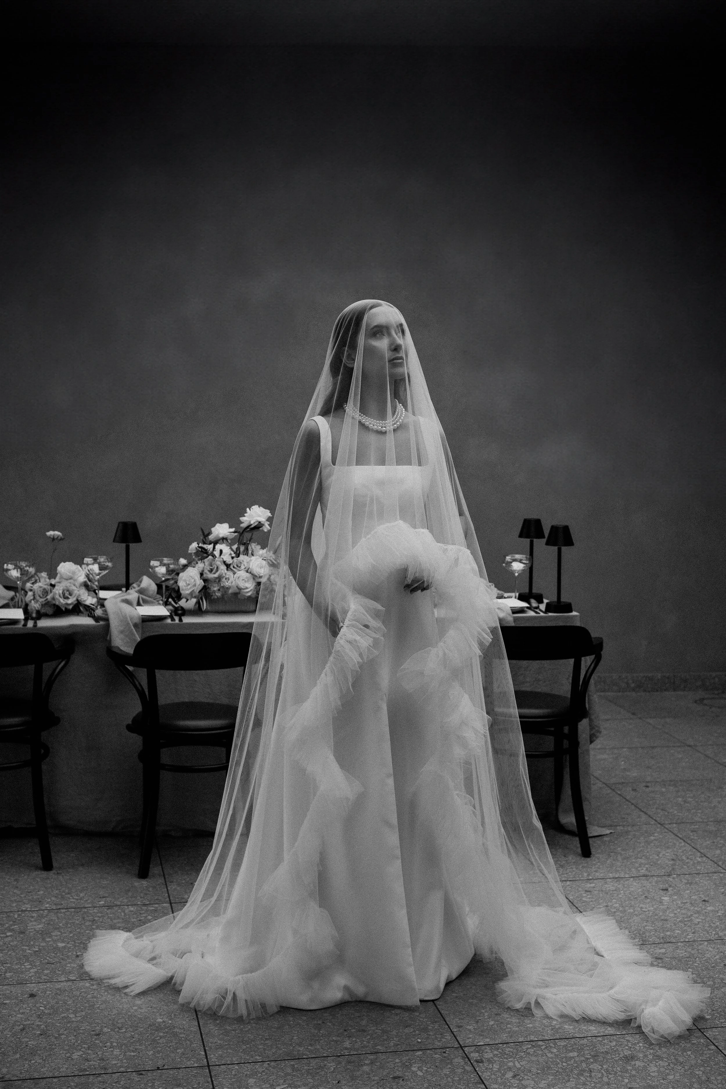 A woman dressed in a wedding gown and veil, holding a bouquet, standing in front of a decorated table with flowers, lamps, and glasses, indoors.