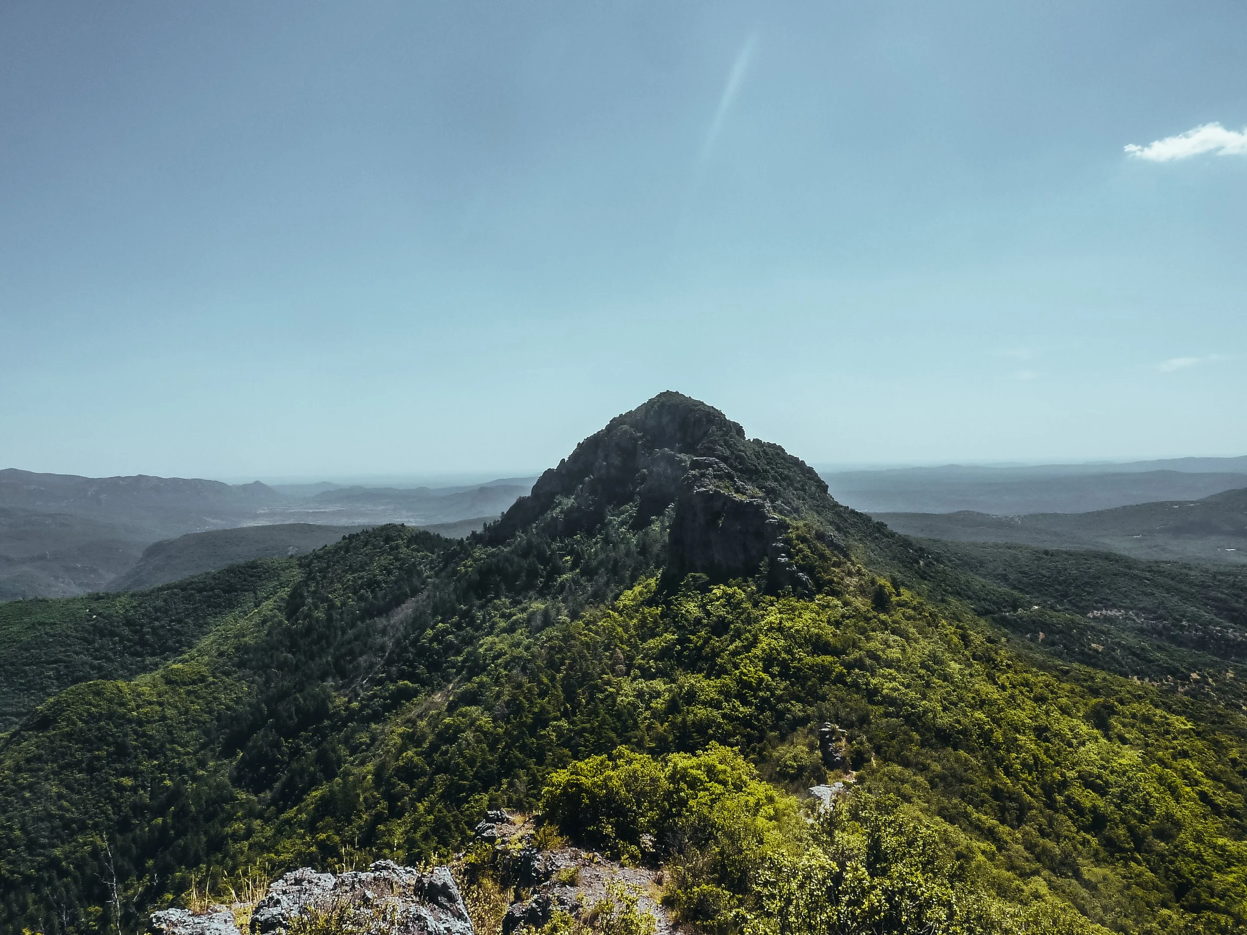 Une vue panoramique d'une montagne rocheuse au sommet entourée de forêts verdoyantes, avec un ciel clair et quelques nuages.