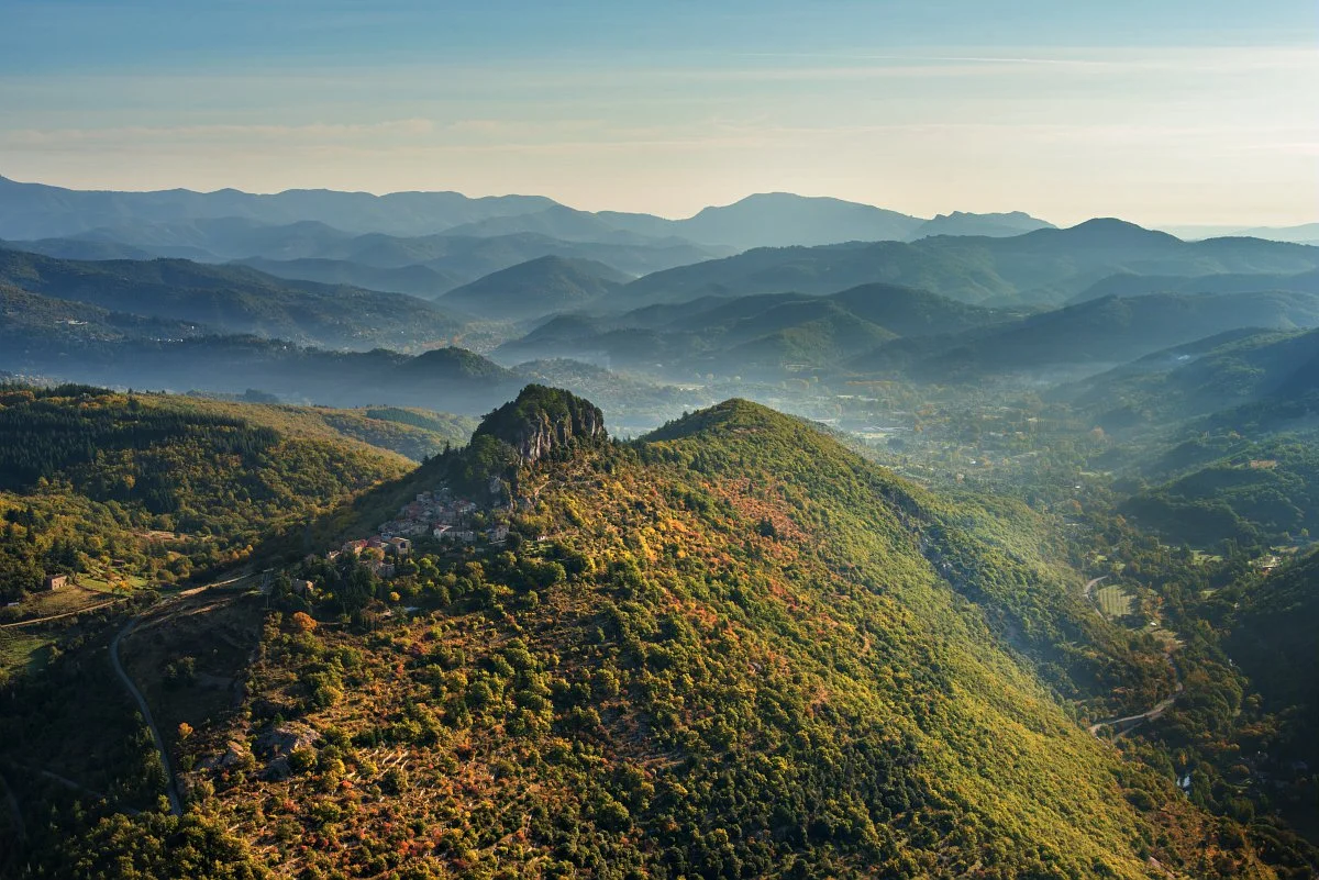 Paysage de montagnes avec une forêt dense, une petite ville accrochée à une colline, et un ciel clair.