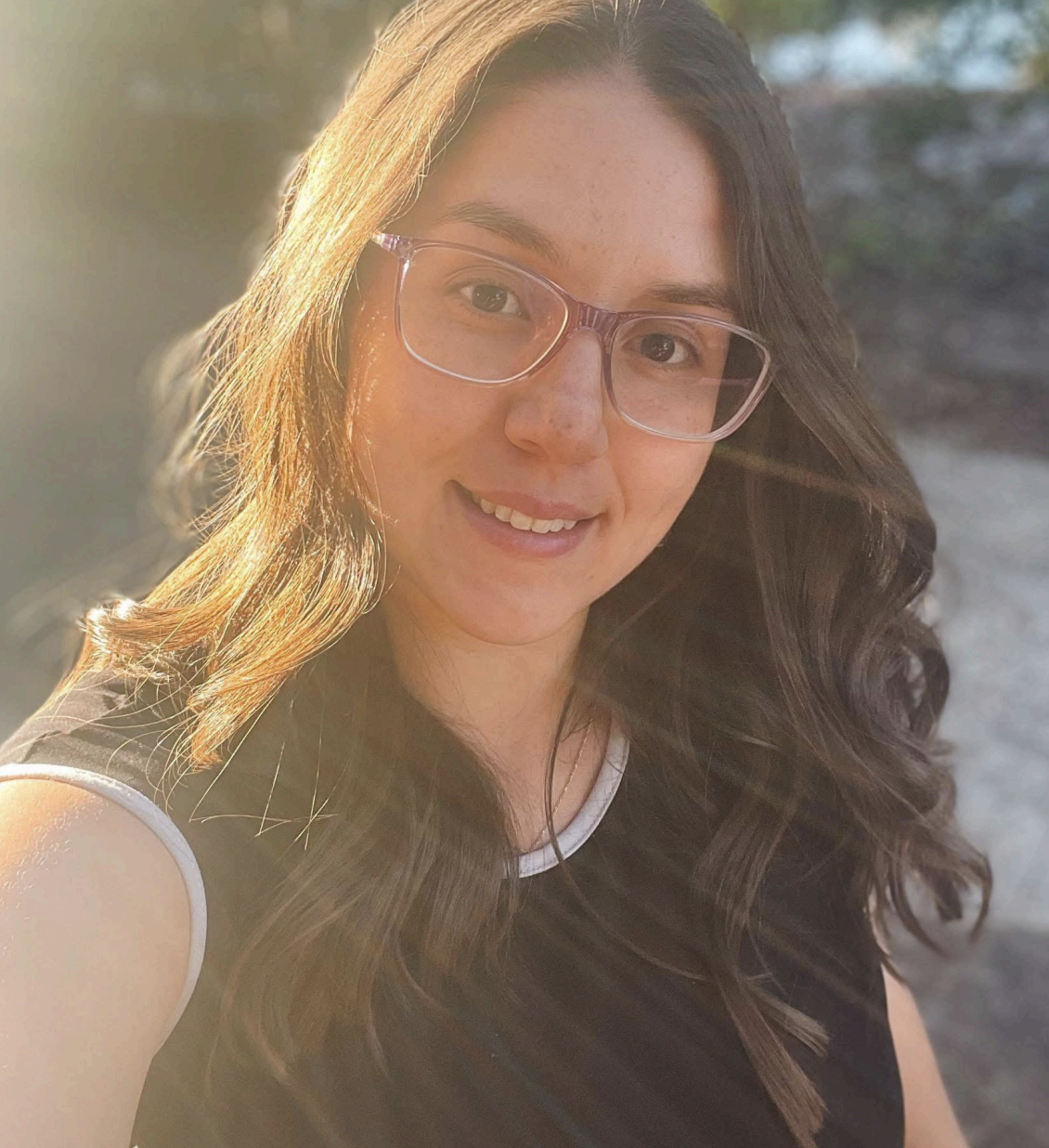 A young woman with long, wavy brown hair wearing glasses, smiling, outdoors during golden hour with sunlight shining on her face.