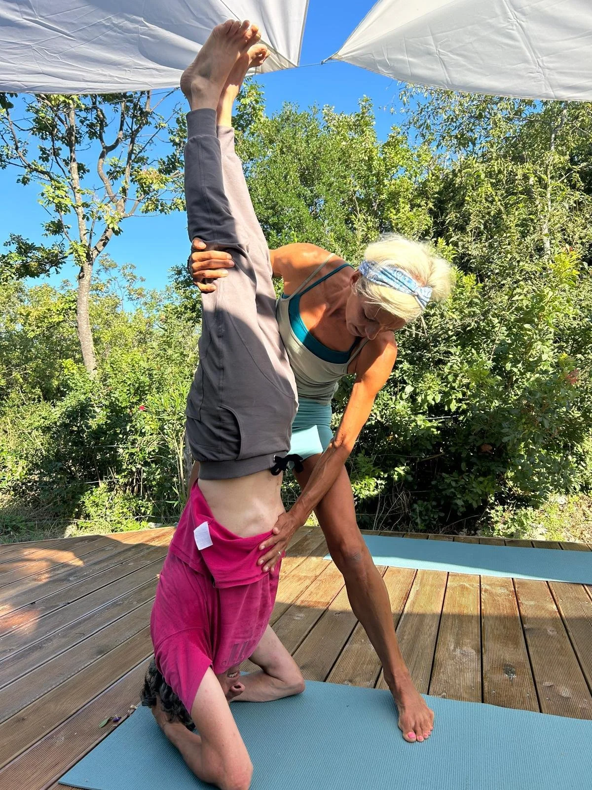 An elderly woman practicing yoga outdoors on a wooden deck, assisting a young girl with a yoga pose. The background features green trees and a bright blue sky.