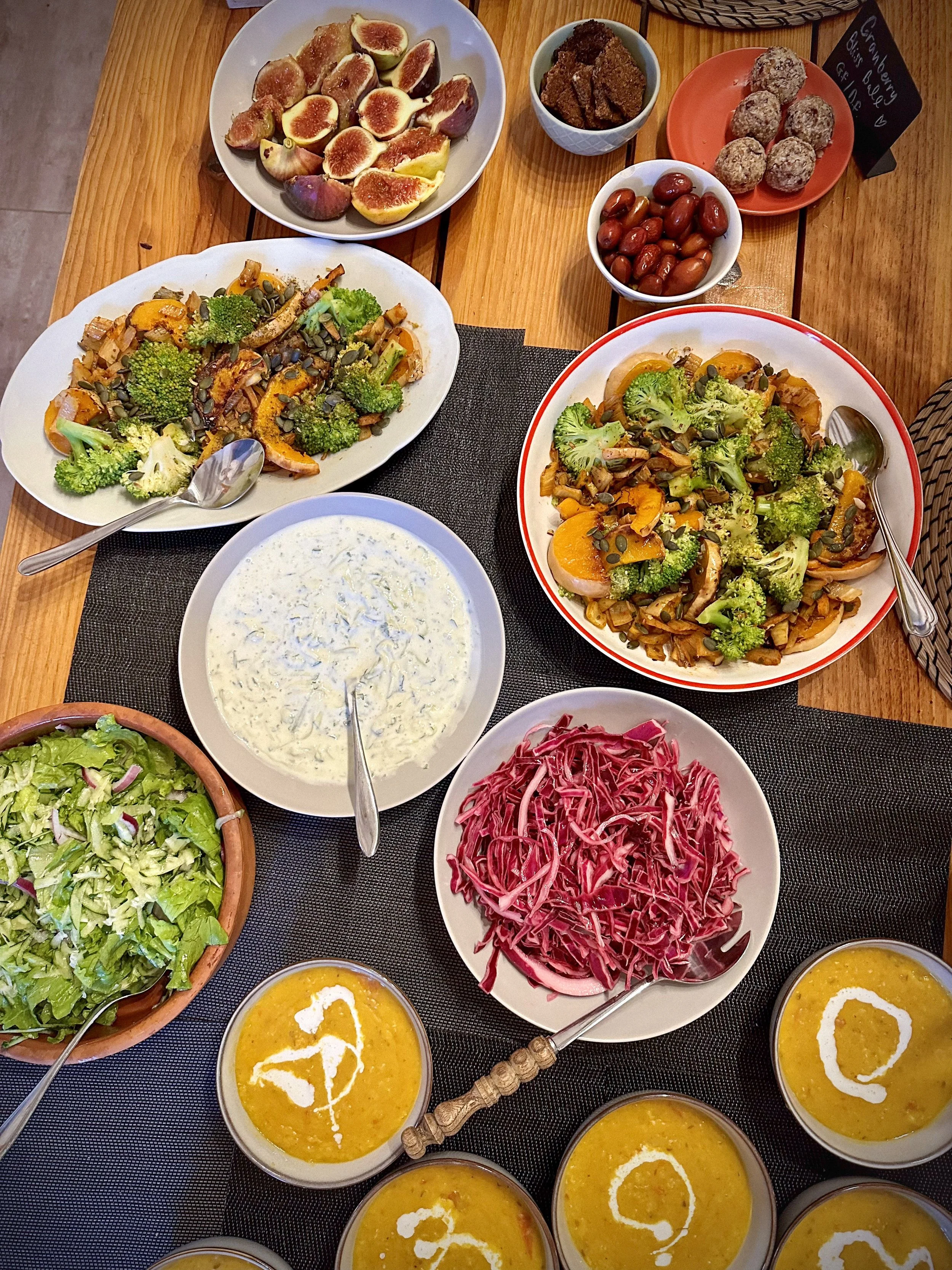 A table filled with various colorful dishes including bowls of creamy orange soup garnished with a dollop of cream, shredded purple cabbage, a fresh green salad, two serving spoons, and plates of roasted vegetables such as broccoli and bell peppers, along with fresh figs, grapes, and desserts like chocolate squares and energy balls.