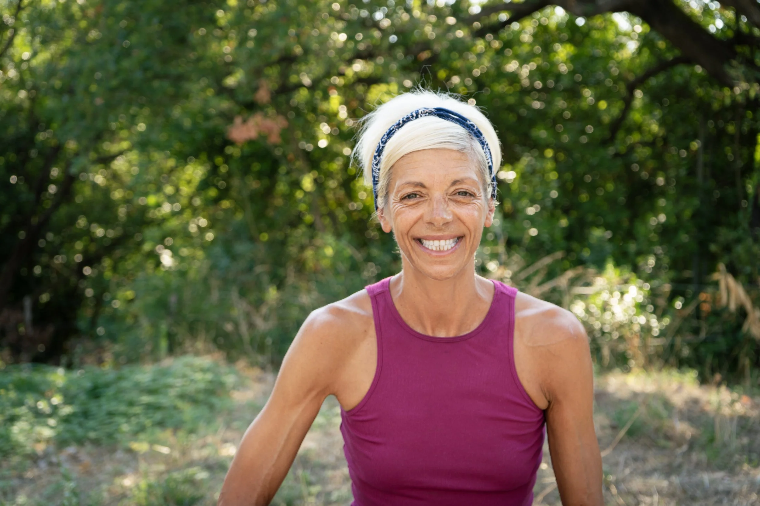 Smiling woman with light blonde hair wearing a purple tank top outdoors on a sunny day with green trees in the background.