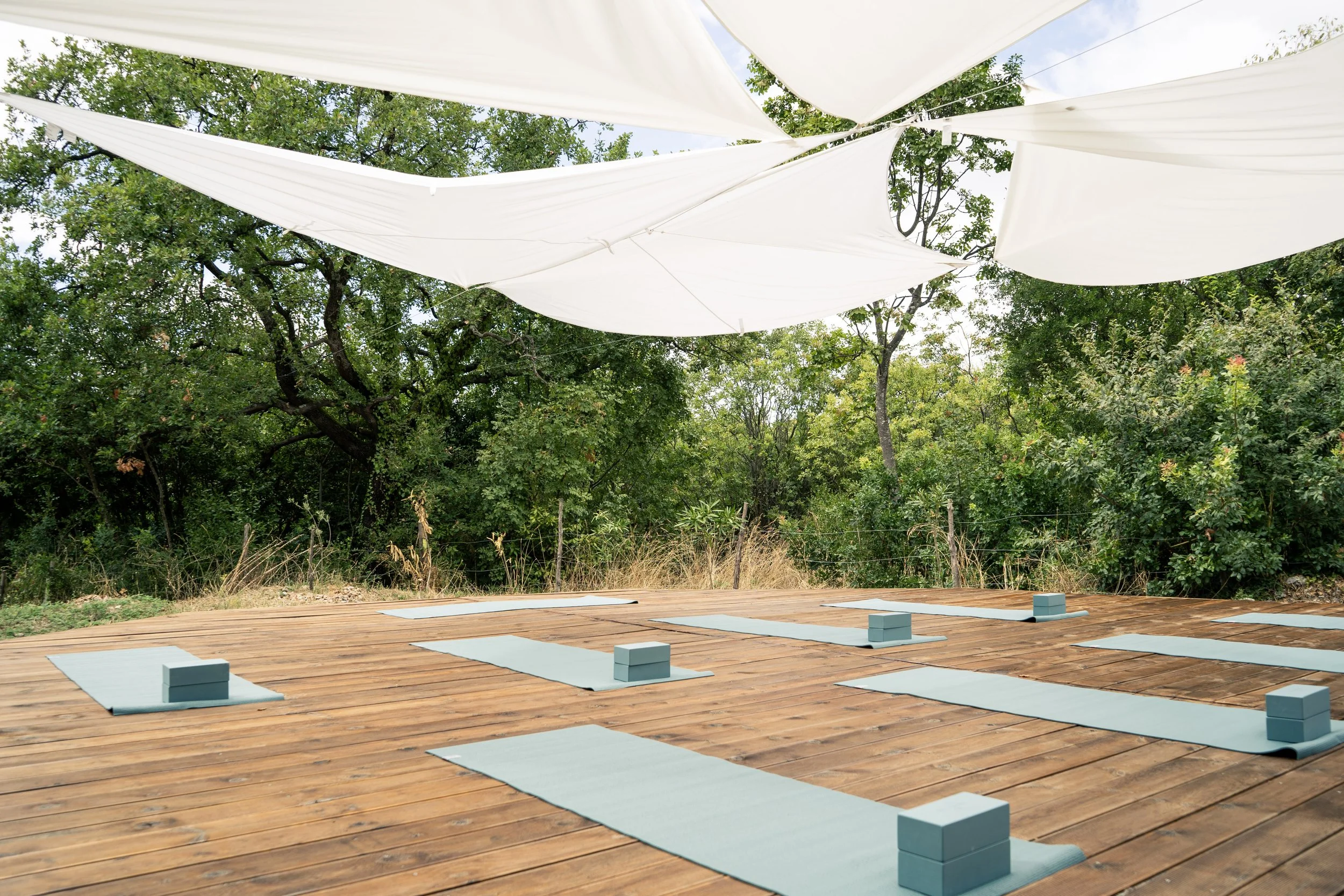 Outdoor yoga setup on a wooden deck with light blue mats and blocks, shaded by large white fabric canopies with trees in the background.
