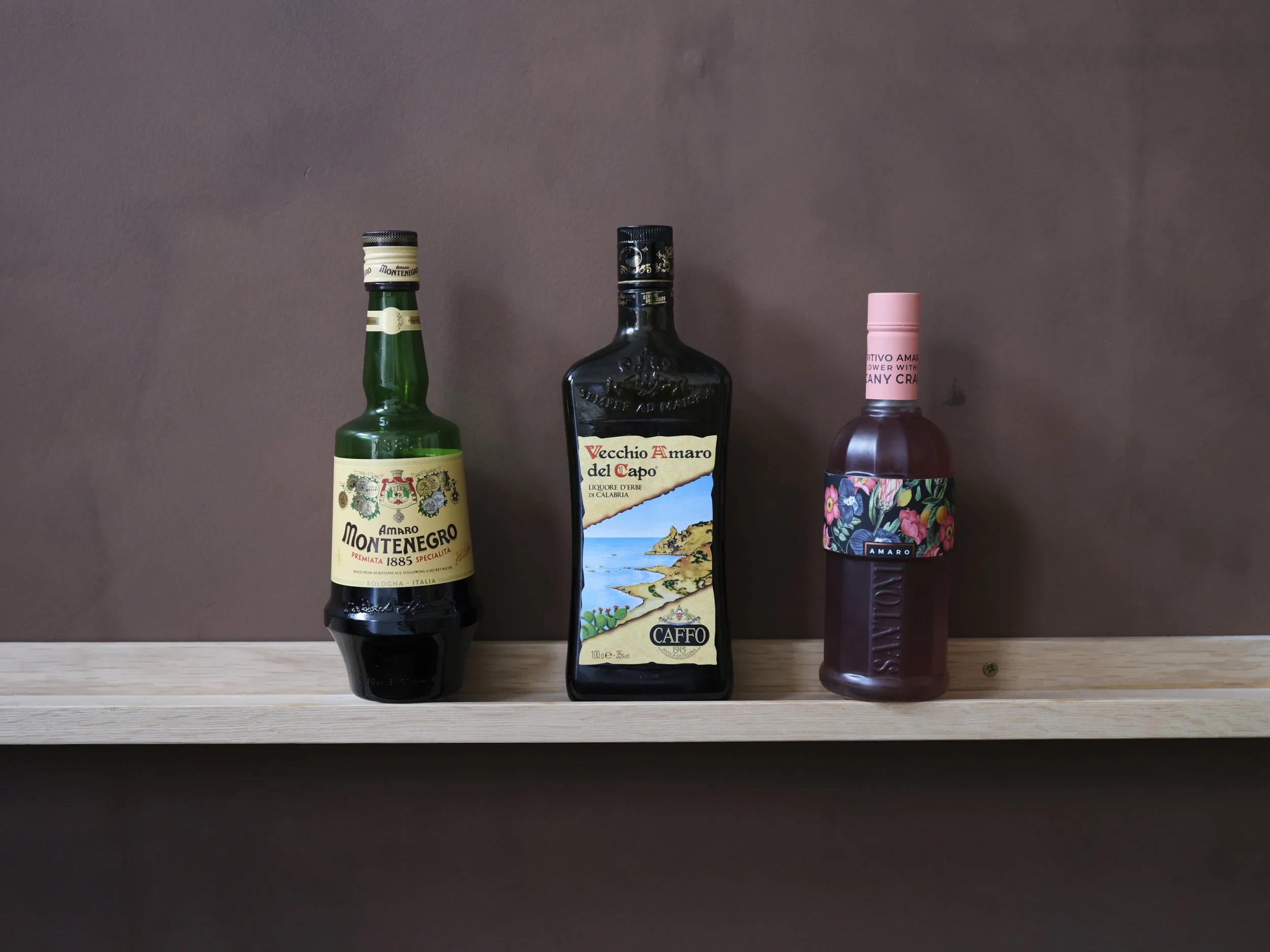 Three bottles of alcohol on a wooden shelf against a plain wall. The first is a green bottle of Montenegro Amaro, the second a black bottle of Vecchio Amaro del Capo, and the third a pink-labeled bottle of floral-flavored liqueur.
