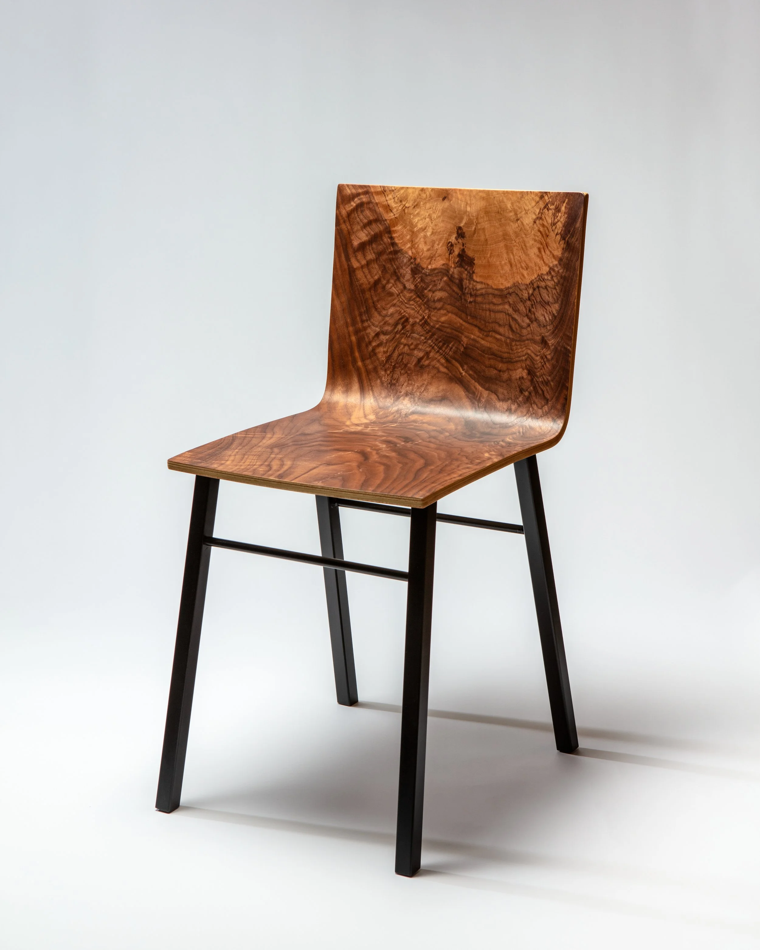 A wooden chair with a curved seat and backrest, featuring a rich wood grain pattern, and supported by black metal legs against a plain background. Chair made by Forma Custom Furniture.