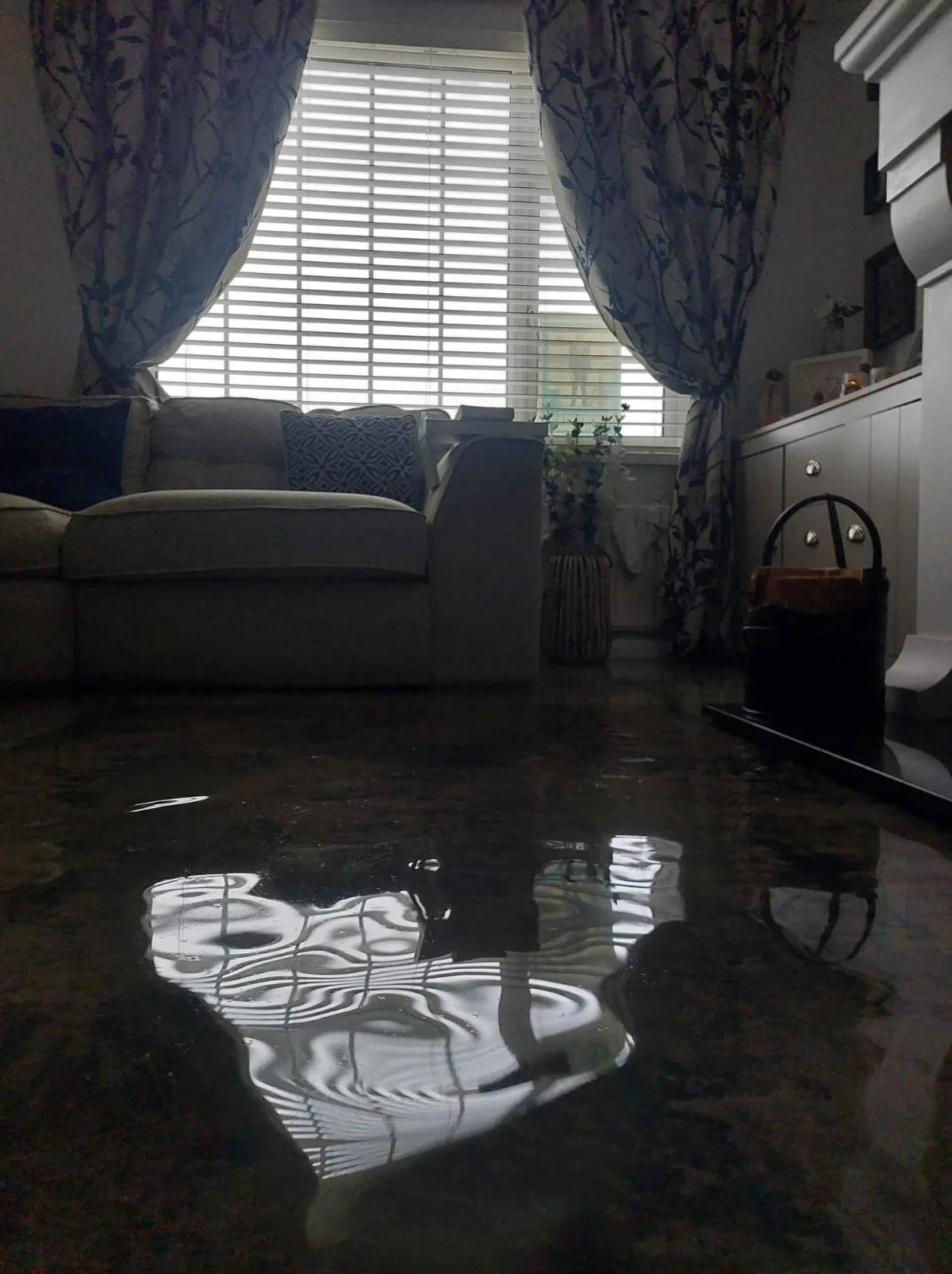 Living room with a floral-patterned curtain framing a window, a beige sofa with decorative pillows, and a table reflecting light from the window. There are also various household items including a cabinet, plants, and a paper towel roll.