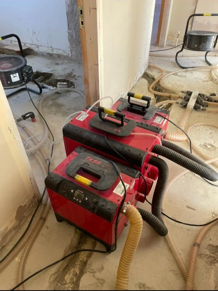 Construction site with two portable dust extractors connected to hoses, surrounded by construction debris and piping in an unfinished room.