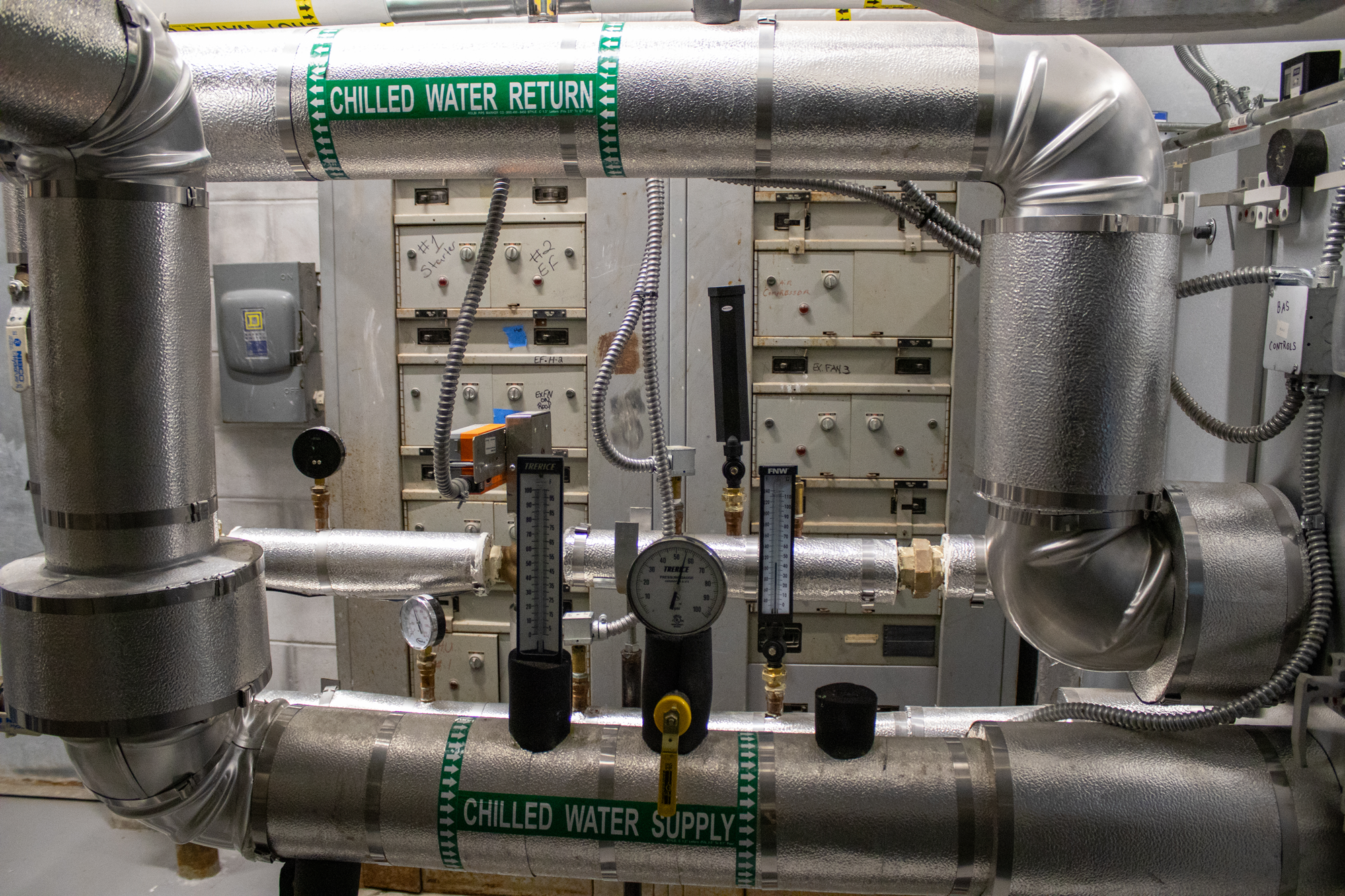 An industrial piping system with insulated silver-colored pipes labeled "Chilled Water Return" and "Chilled Water Supply". The system includes gauges, valves, and control panels for managing water flow.