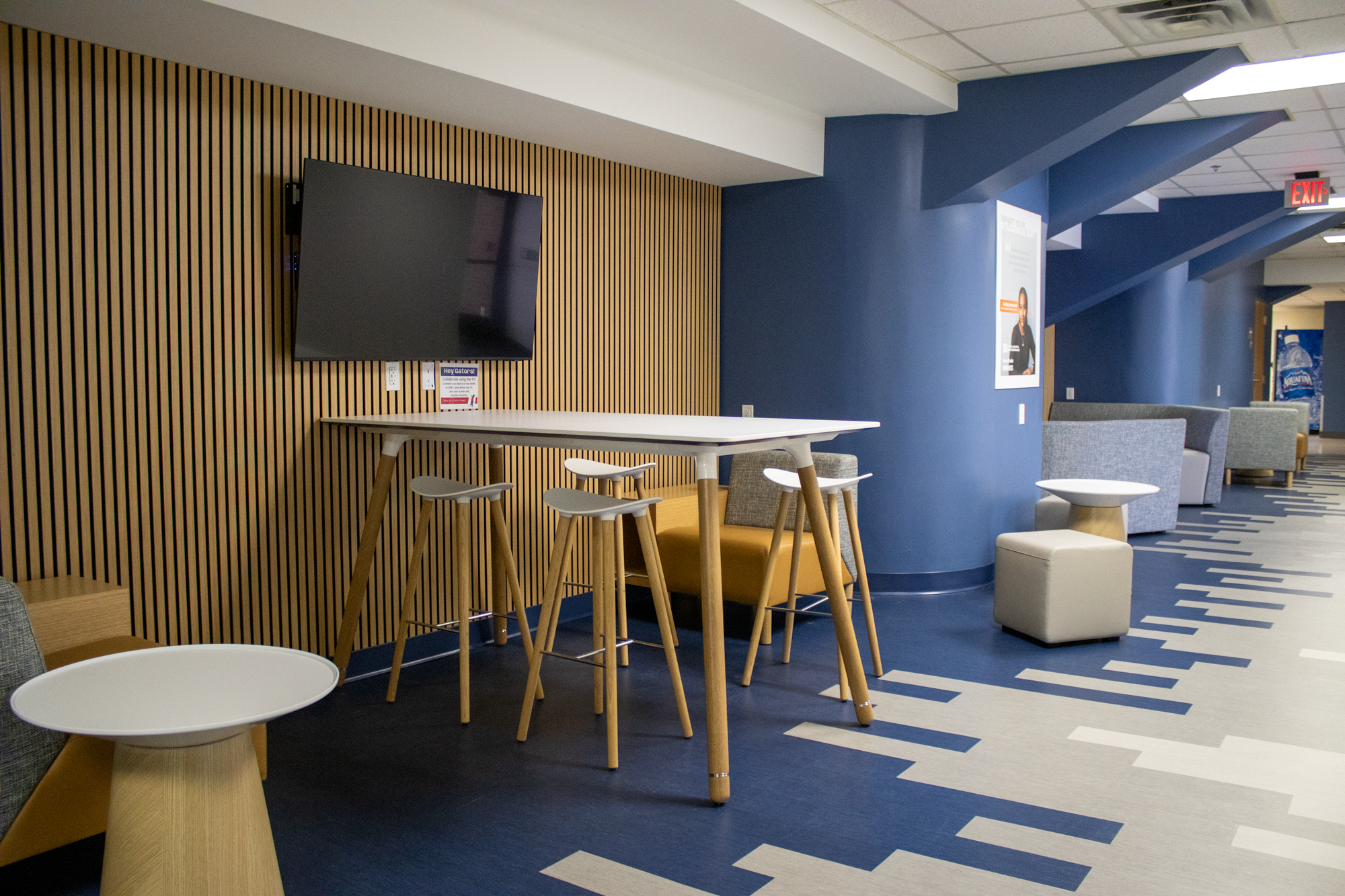 Modern lounge area with high table and stools, gray and mustard chairs, a wall-mounted TV, and a blue and gray patterned carpet.
