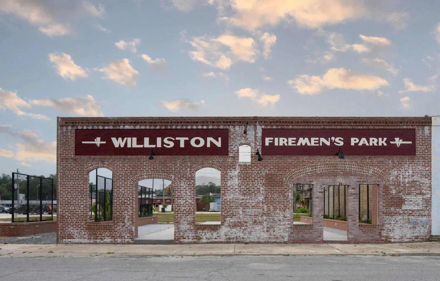 A brick wall with signs reading 'Williston Firemen's Park' and three arched openings, set against a partly cloudy sky.