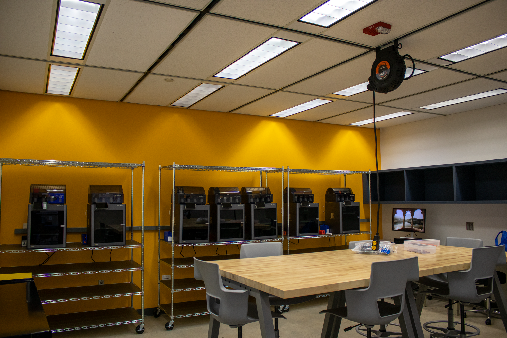A room with a large wooden table surrounded by chairs, a yellow wall, and a shelving unit with several 3D printers. There is a small monitor on the right side of the room and a ceiling mounted retractable tape measure.