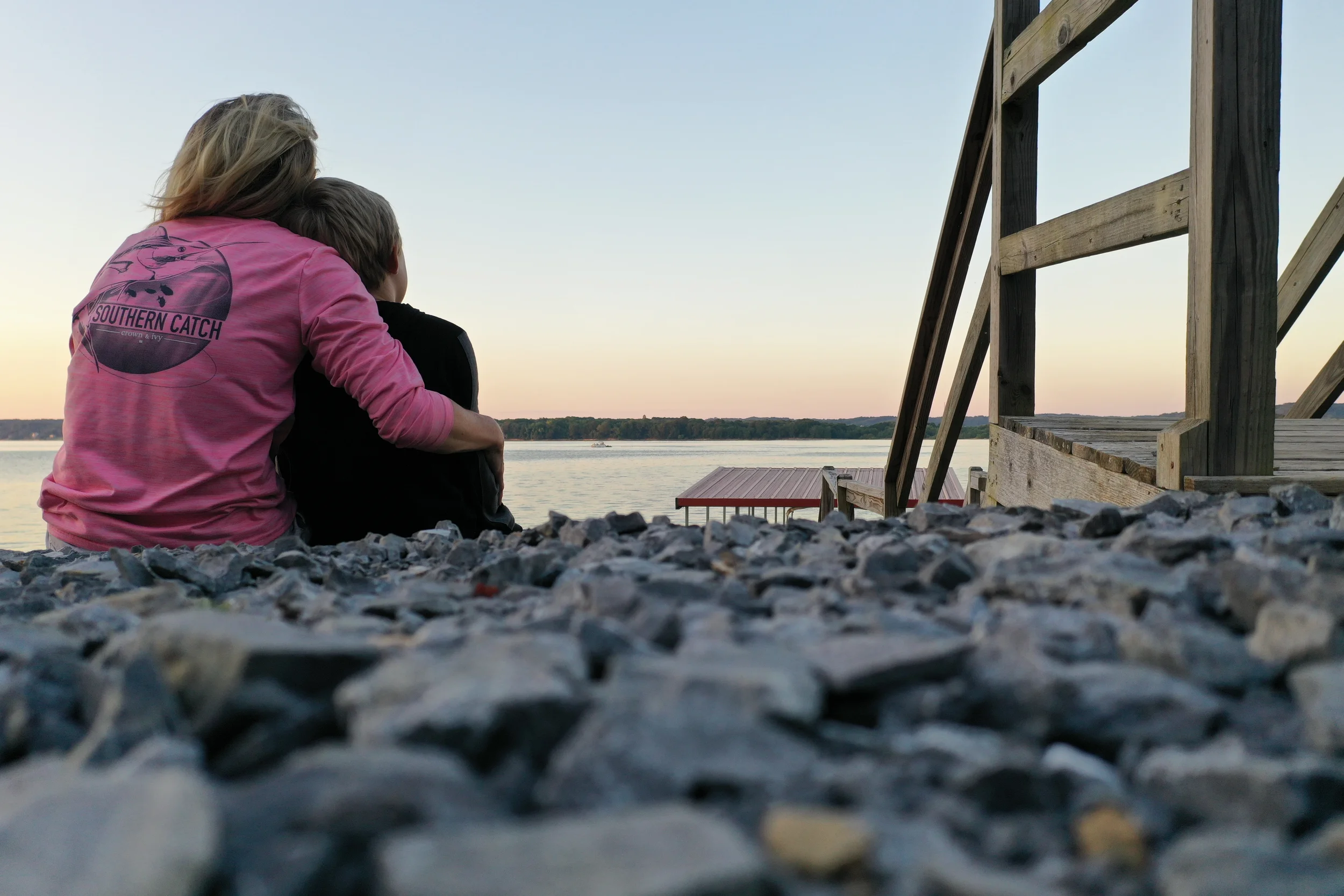 A woman and a boy sitting by a lake at sunset, facing the water.