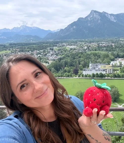 Lauren Haley being silly, holding a strawberry plushy in her hand, posing for a travel selfie