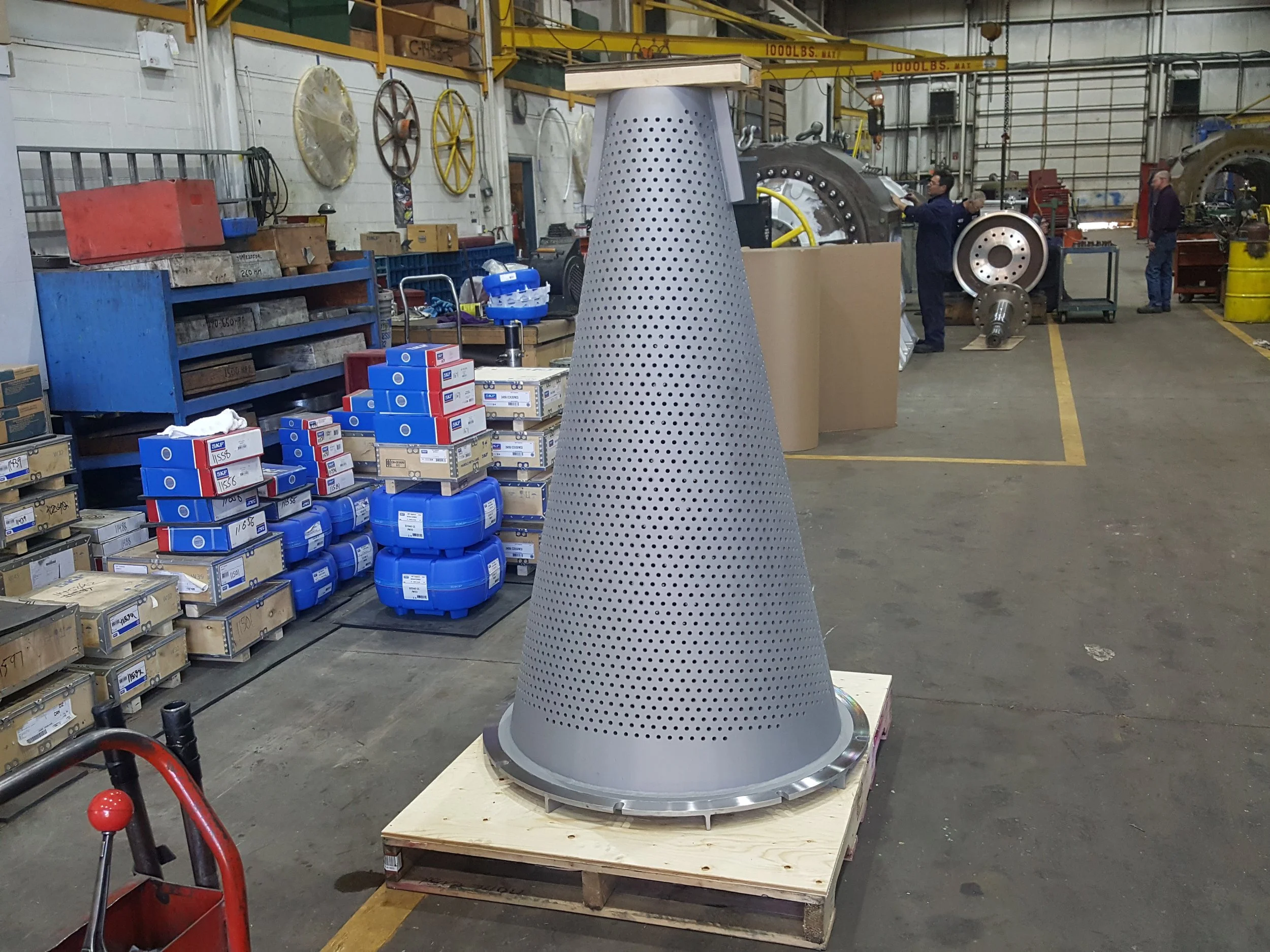 Sunrise Engineering - A large metallic cone with perforations, placed on a wooden pallet inside an industrial workshop. In the background, there are workers, machinery, and shelves with boxes and parts.