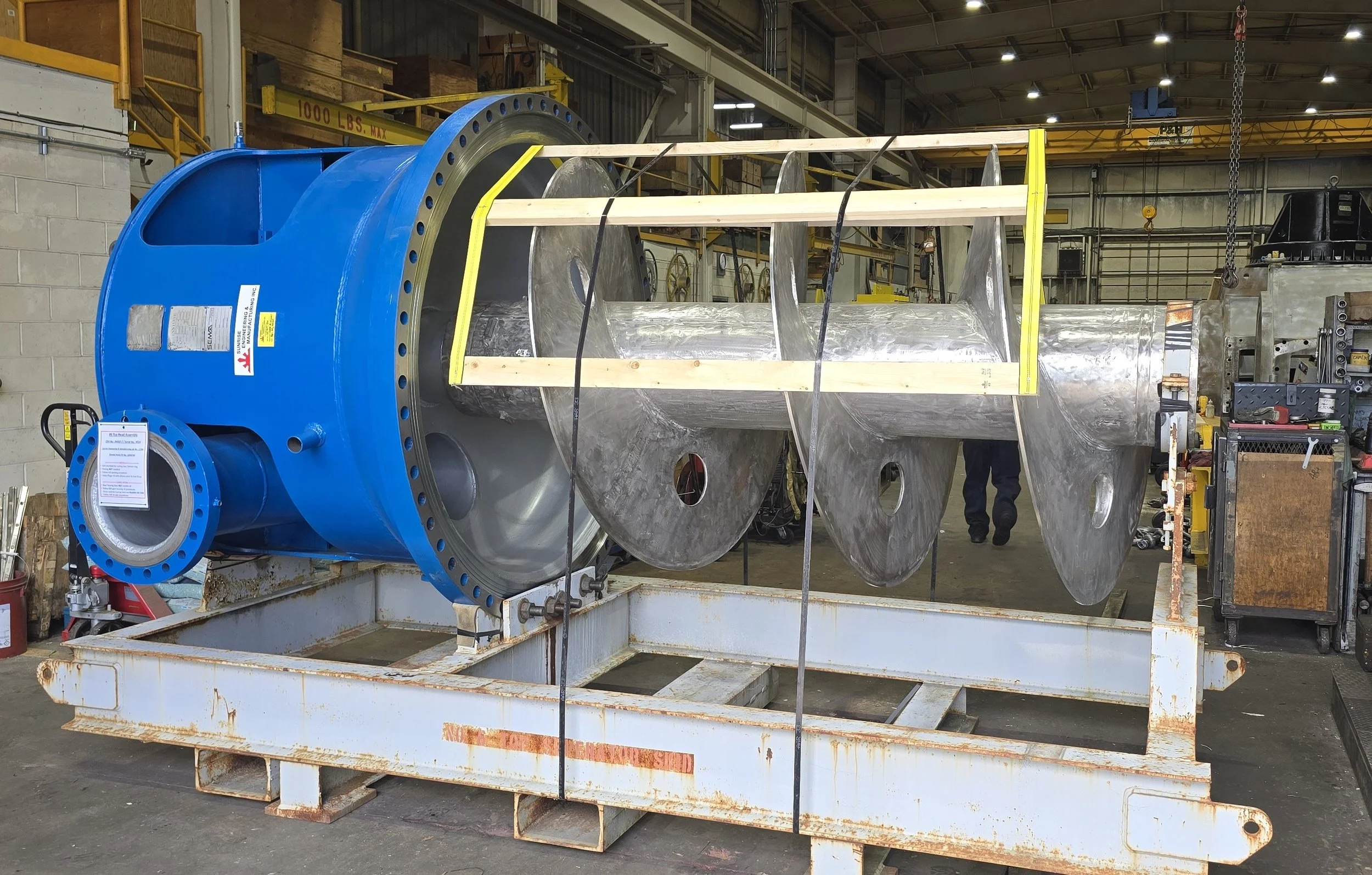 Sunrise Engineering - Large industrial assembly with a blue cylindrical component attached to a metallic structure, supported by a rusty white frame, situated in an industrial warehouse.