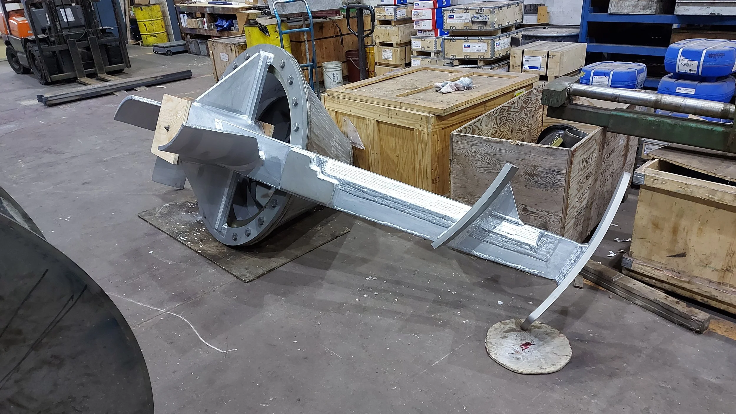 Sunrise Engineering - metallic industrial machine component with a circular end and a long arm, placed on a workshop floor next to wooden crates and storage shelves.