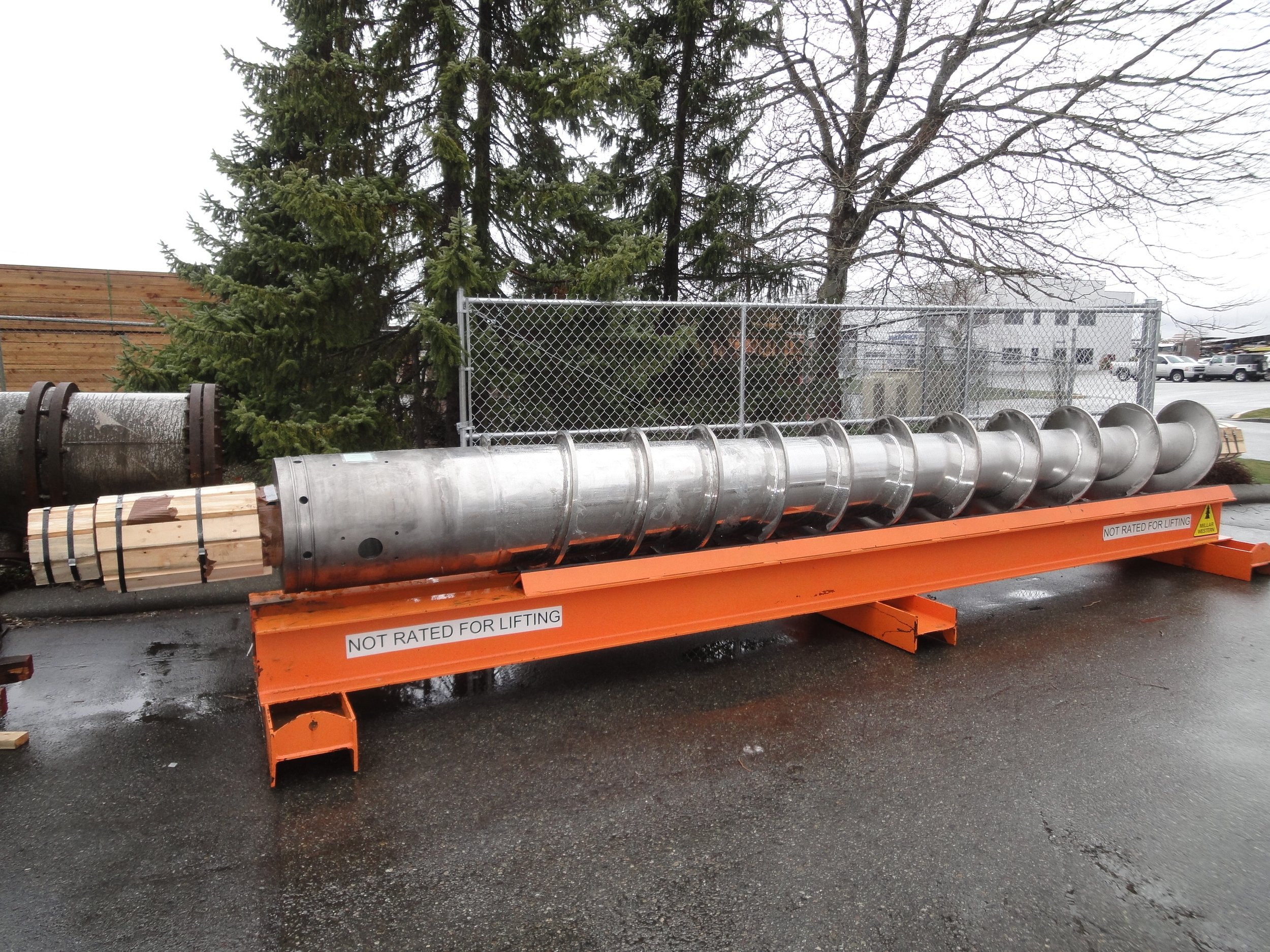 Large metallic screw conveyor with wooden pallets attached, placed on an orange metal platform marked 'Not Rated for Lifting' in an outdoor area with trees, a chain-link fence, and a parking lot in the background.