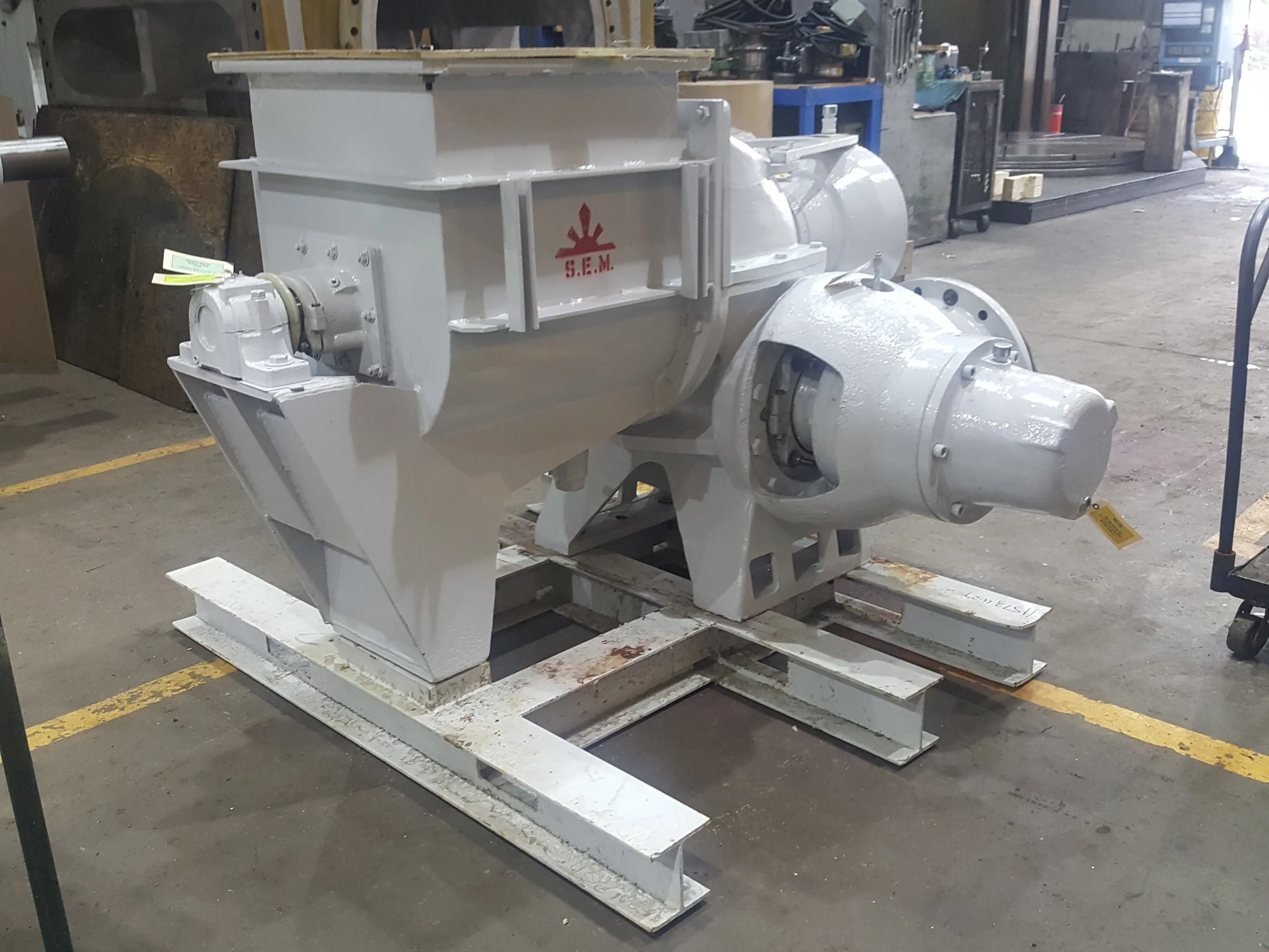 Large industrial piece of equipment painted white with a red logo reading 'S.E.M.' on the side, situated in a workshop or factory setting with tools and machinery in the background.