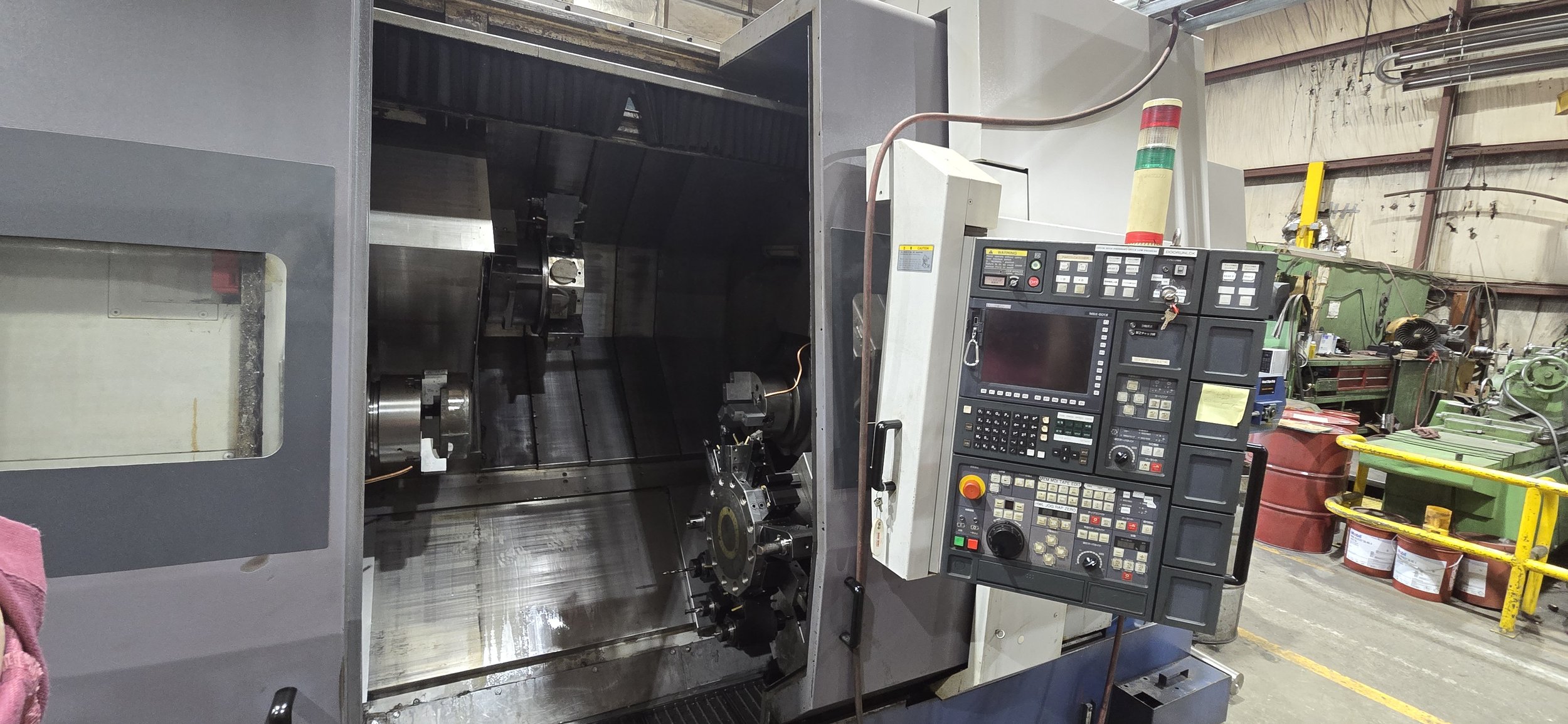 Sunrise Engineering -  large industrial CNC machine, with a control panel with numerous buttons and a screen, in a workshop with various tools, drums, and machinery in the background.