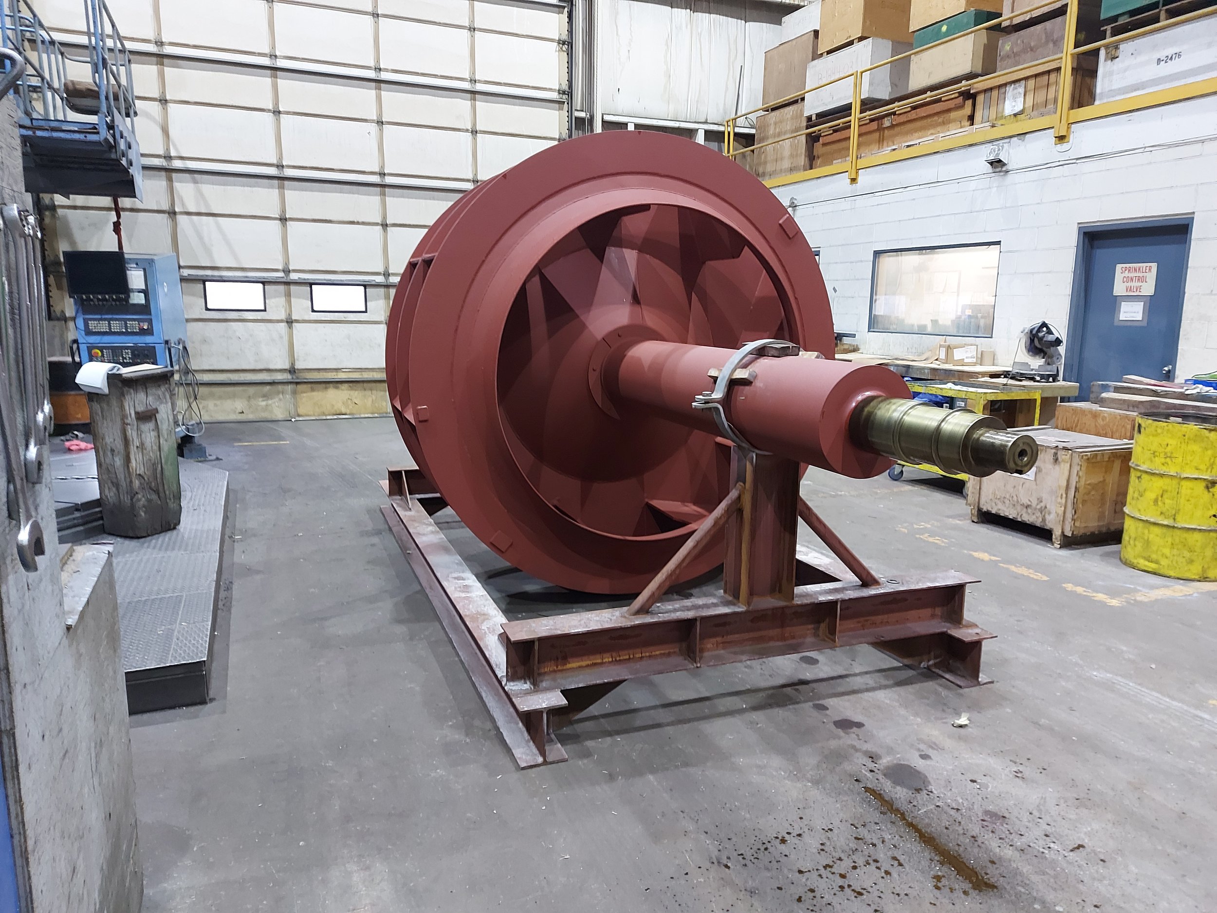 Large industrial turbine or rotor on a metal stand inside a warehouse with equipment and tools around.