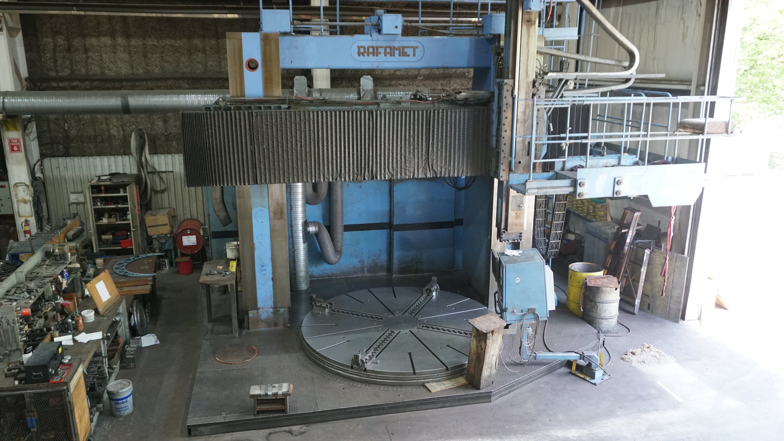 Sunrise Engineering - Industrial workshop with large blue CNC machine labeled 'RAFAMET', metal work surfaces, tools, shelves, and equipment, with a partially open door to the outside.