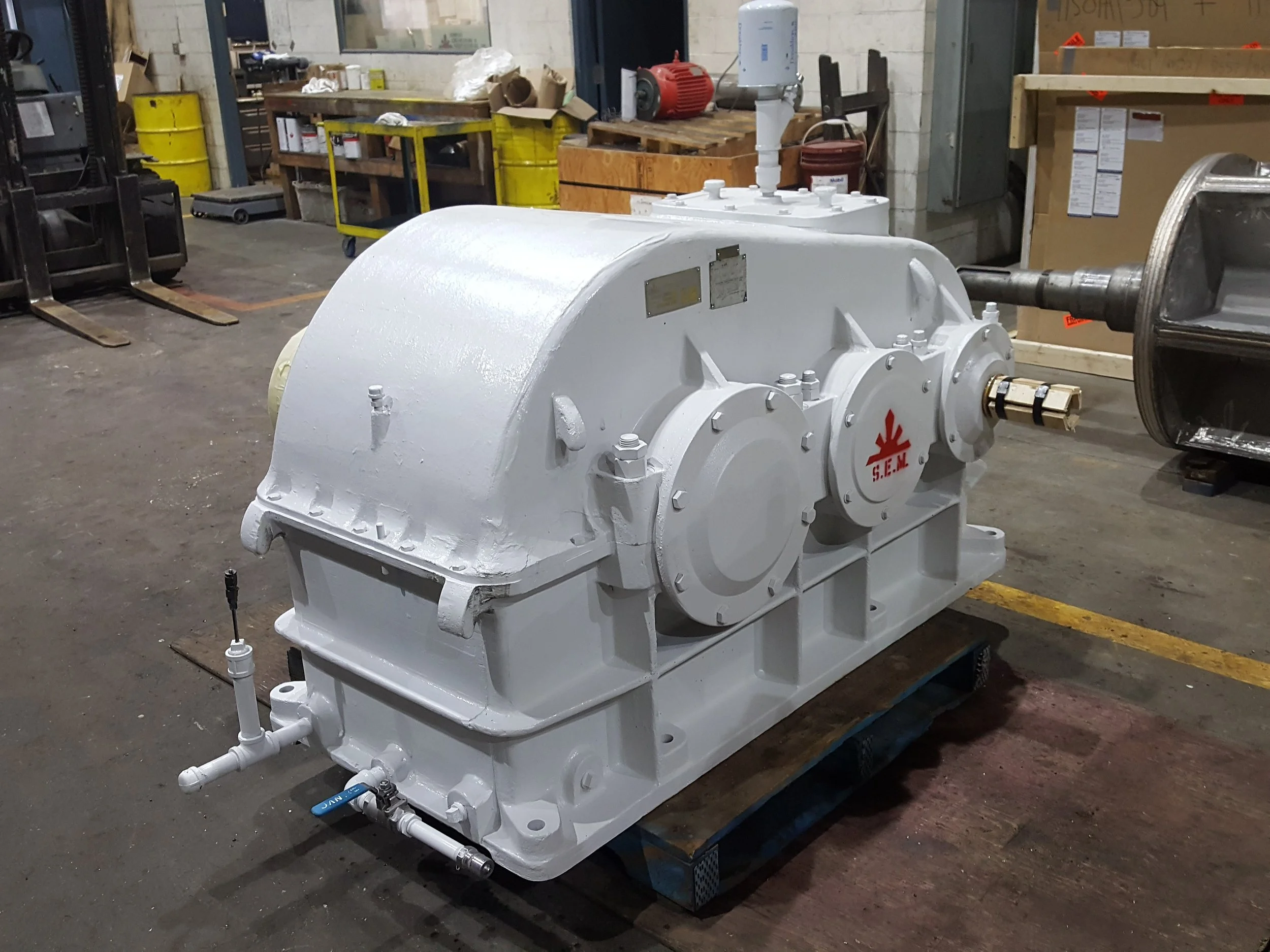 Large industrial gear or gearbox painted white, situated in a factory or workshop environment with tools and storage in the background.
