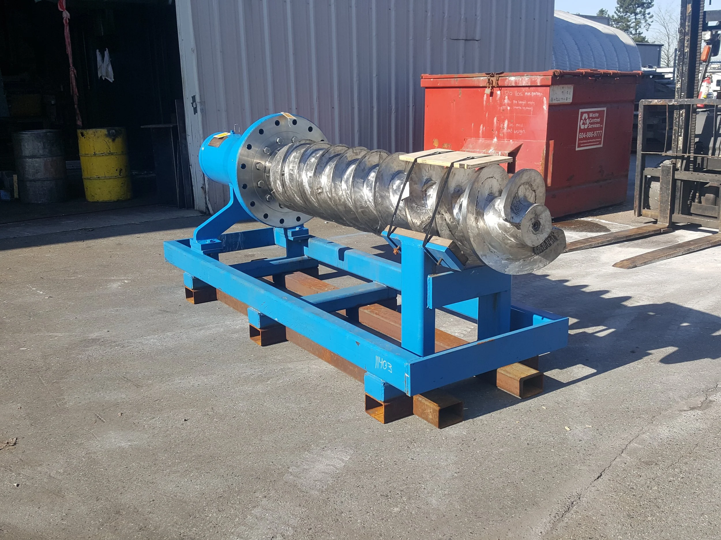 Sunrise Engineering - metal industrial screw or auger mounted on a blue stand outside near a warehouse with yellow barrels and a red dumpster.