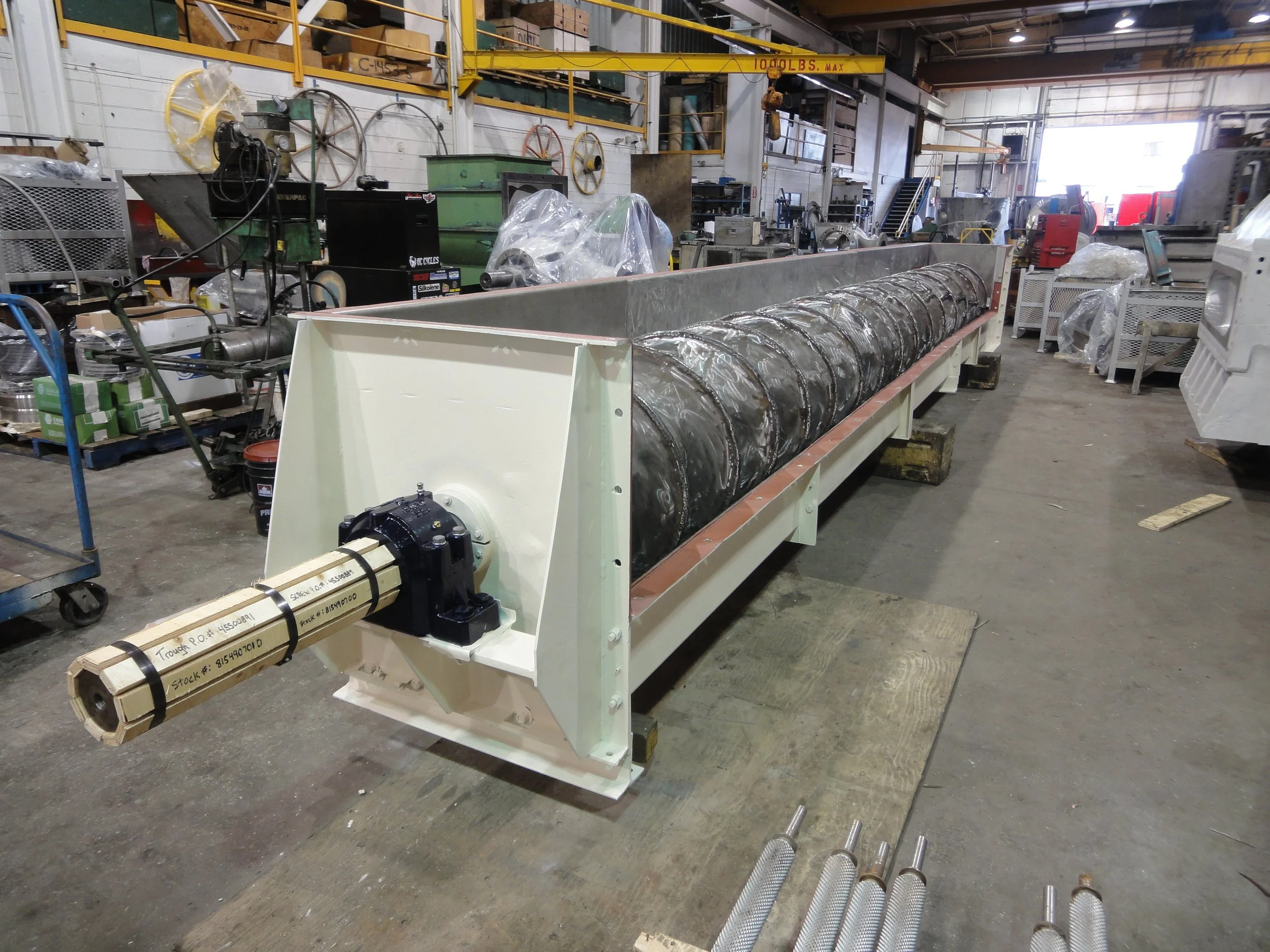 Sunrise Engineering -  a long cylindrical component, situated in a workshop or factory setting. The equipment is mounted on a white frame and has a shaft at one end. The background includes shelves, tools, and various machinery.