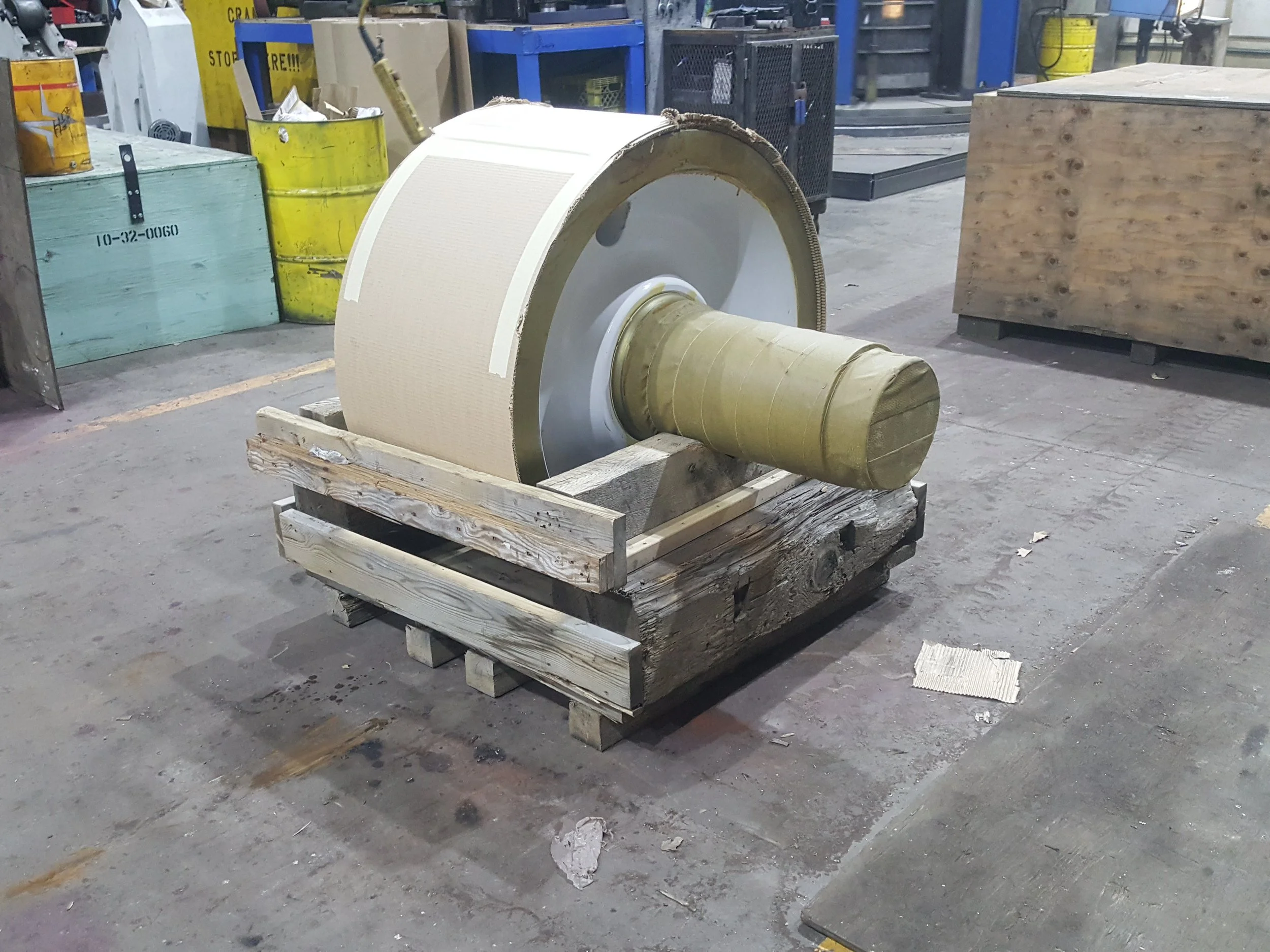 Large industrial engine part with a beige outer shell and a gold-colored exhaust pipe, mounted on a wooden pallet in a factory or workshop setting.