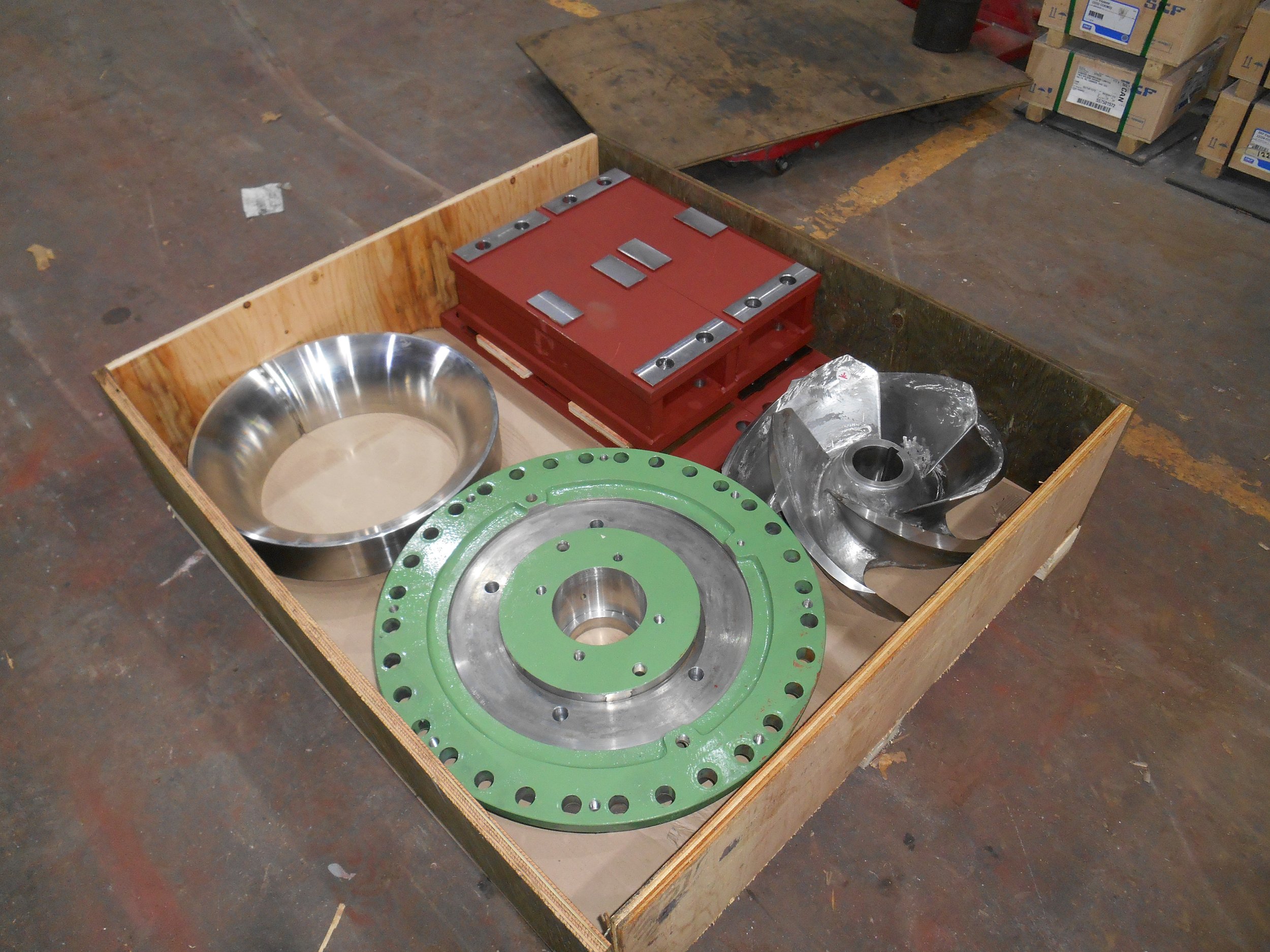 Sunrise Engineering - Various metal machine parts in a wooden crate on a workshop floor, including a large green and silver gear, a shiny metal bowl, and other metallic components.