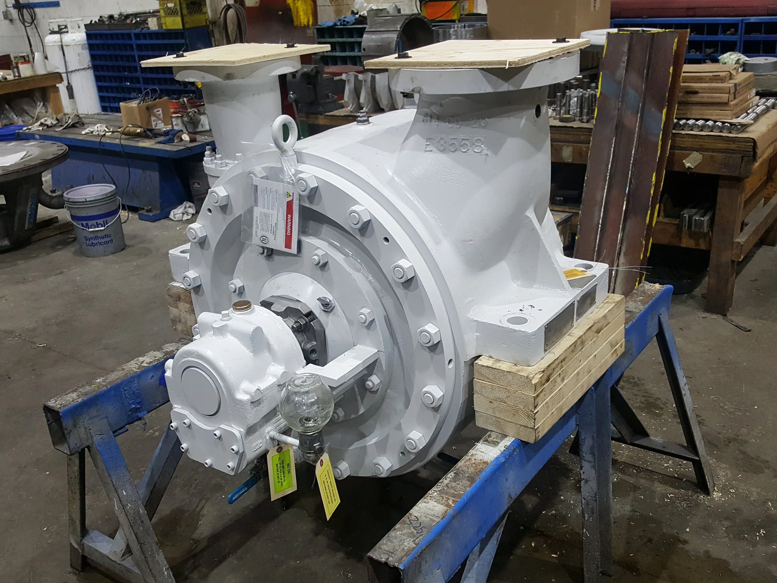 Large white industrial pump or gearbox mounted on a workbench in a workshop, surrounded by tools and equipment.