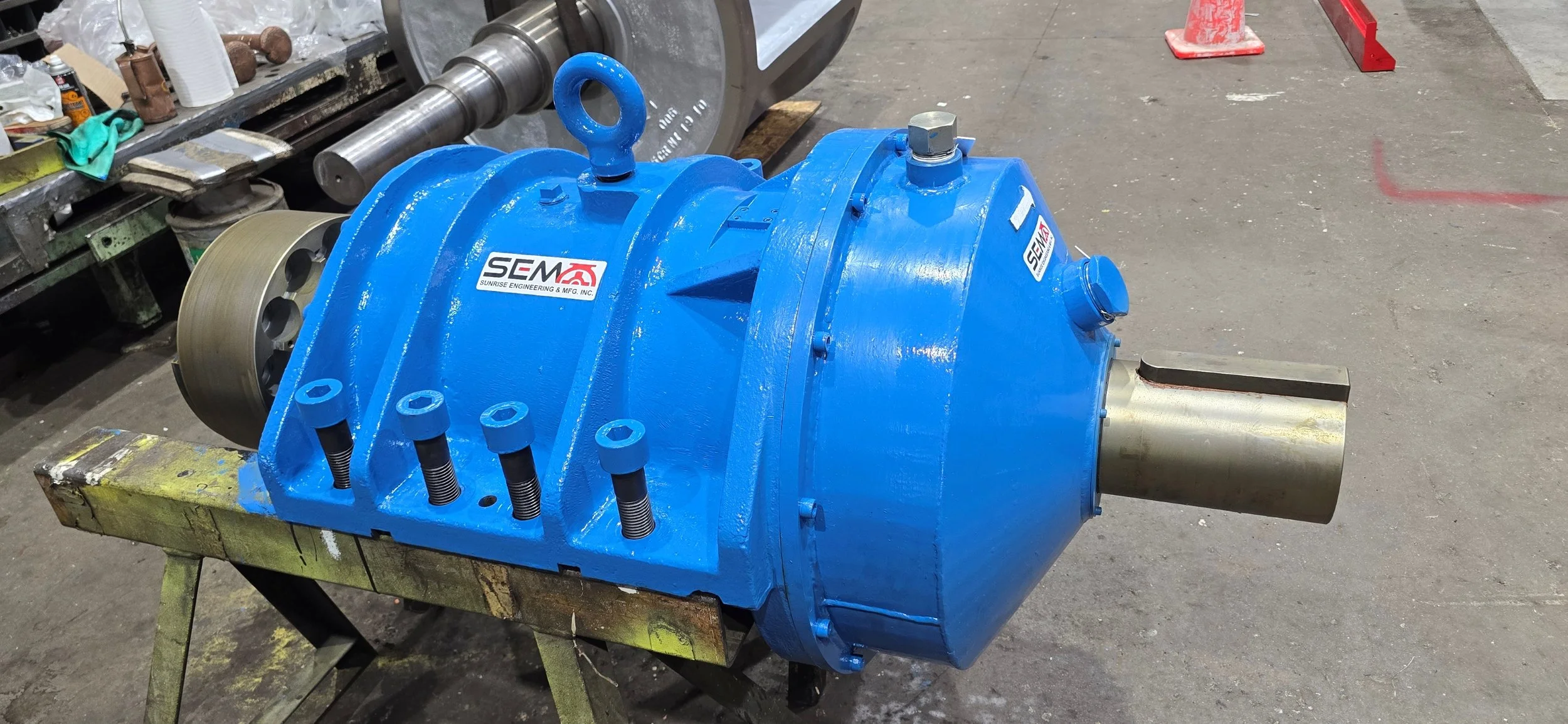 Sunrise Engineering -  A large industrial blue pump or motor part with a metal shaft extending outward, placed on a workbench in a workshop. The part has a SEM logo sticker on it, with various tools and equipment visible in the background.