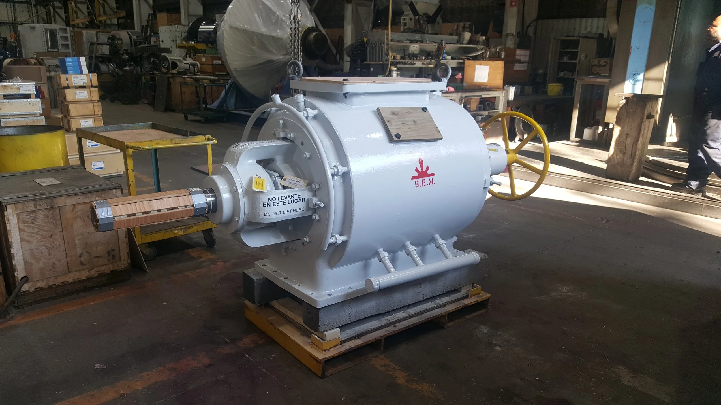 Sunrise Engineering - Large industrial machine painted white with a yellow handwheel, mounted on a wooden pallet in a workshop, surrounded by tools and equipment.
