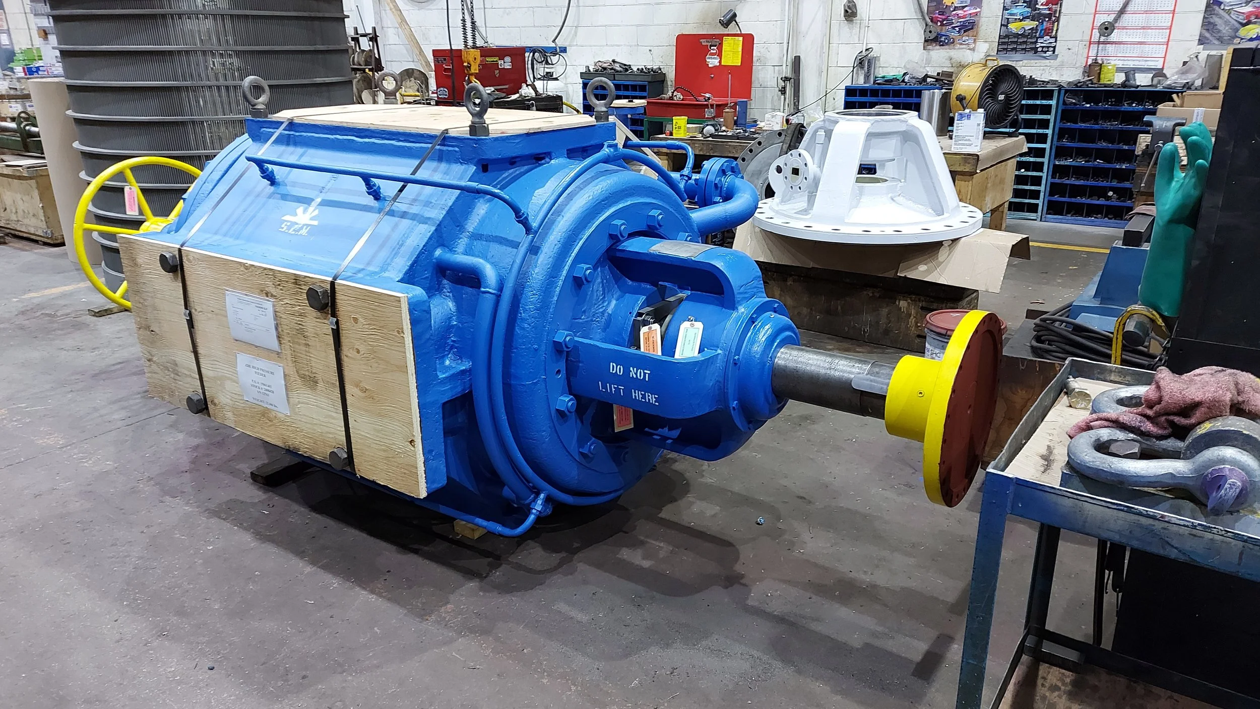 Sunrise Engineering - industrial machine with a cylindrical shape and a shaft protruding from the front, marked with a sign that says 'Do Not Lift Here', placed on a wooden pallet inside a workshop, with various tools and equipment surrounding it.