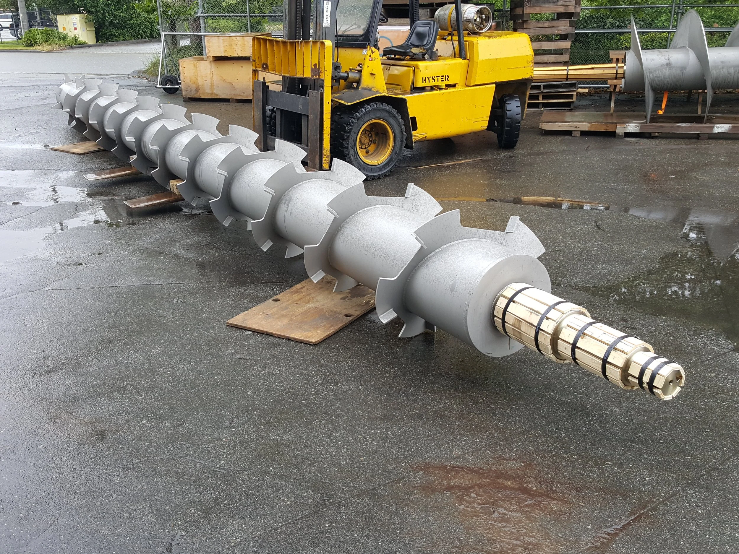 Sunrise Engineering - industrial auger drill bit lying on the ground next to a yellow forklift, with wet pavement and some industrial equipment in the background.