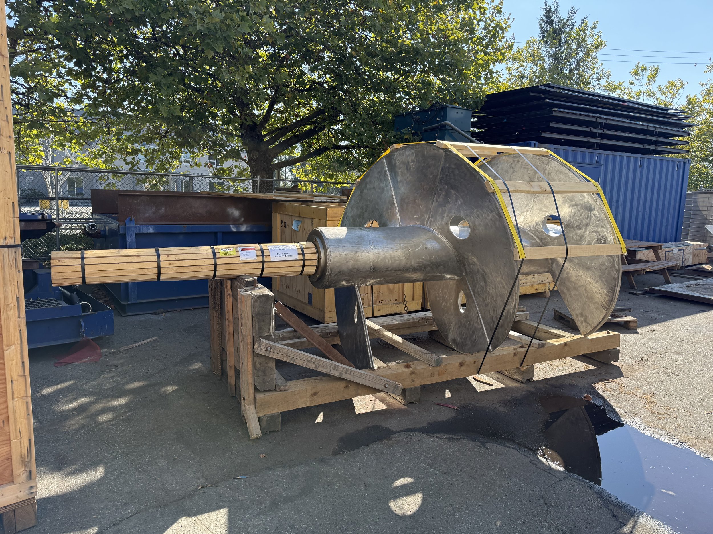 Sunrise Engineering - Large metal object with a wooden handle, resting on a wooden pallet outdoors, surrounded by trees and storage containers.