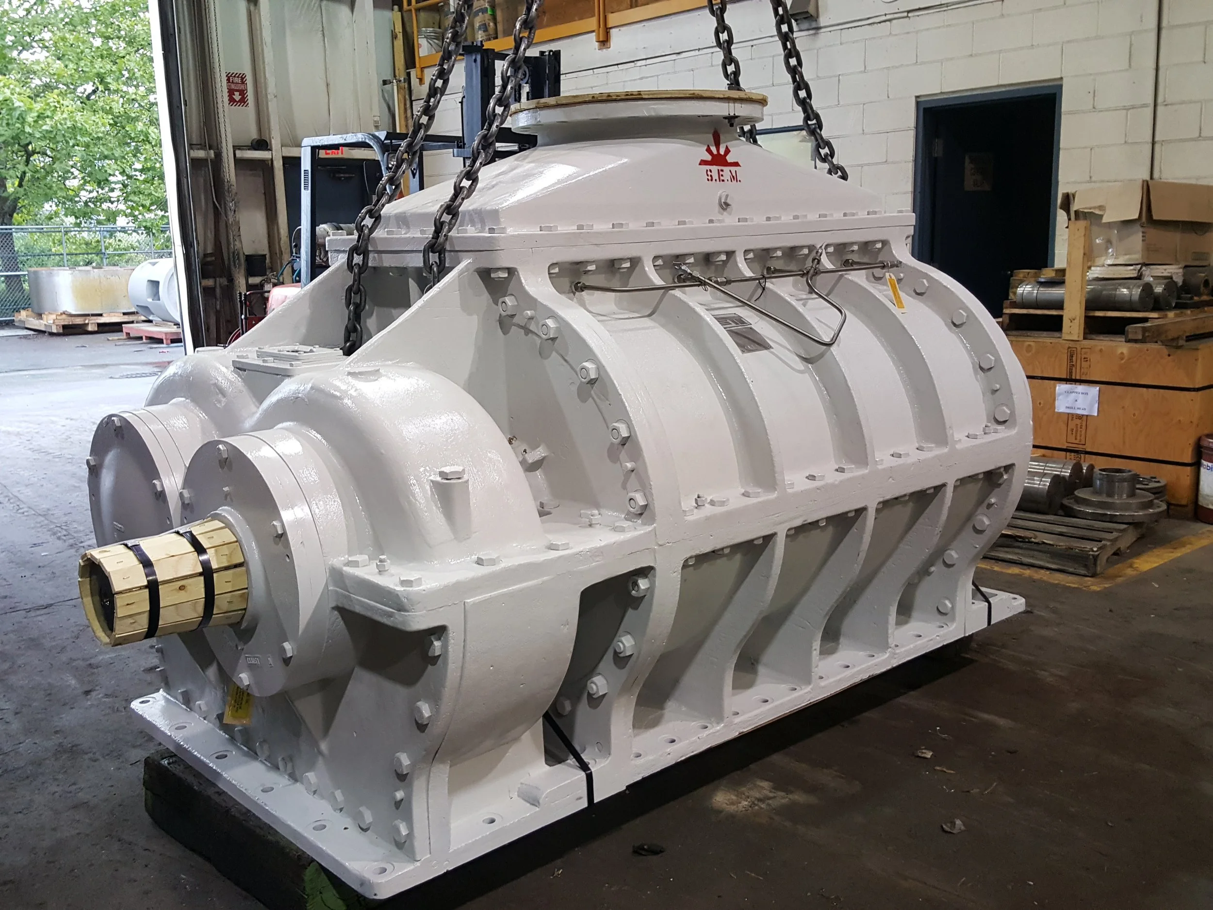 Large industrial machine painted white, with a bolted metal casing, multiple bolts, and a wooden splined shaft at one end, situated in a warehouse.