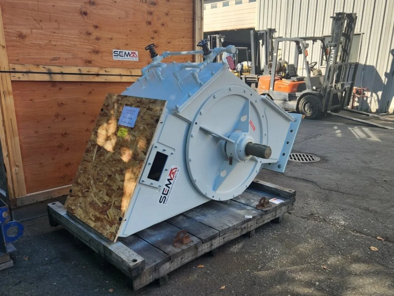 Sunrise Engineering - Large industrial white machinery with attached pipes and a rotating shaft, sitting on a wooden pallet outdoors, with forklifts and a wooden wall in the background.