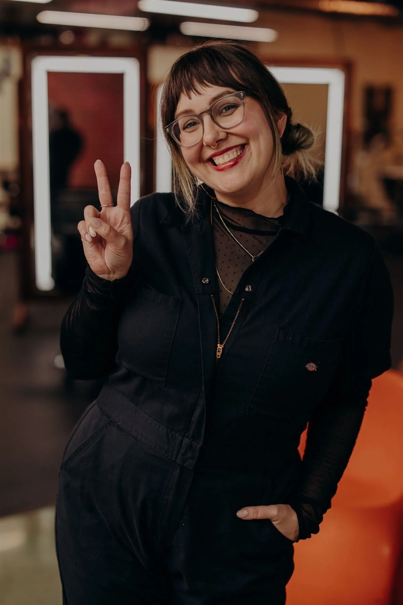 A young woman with blonde hair, glasses, and tattoos, smiling in front of a purple background.