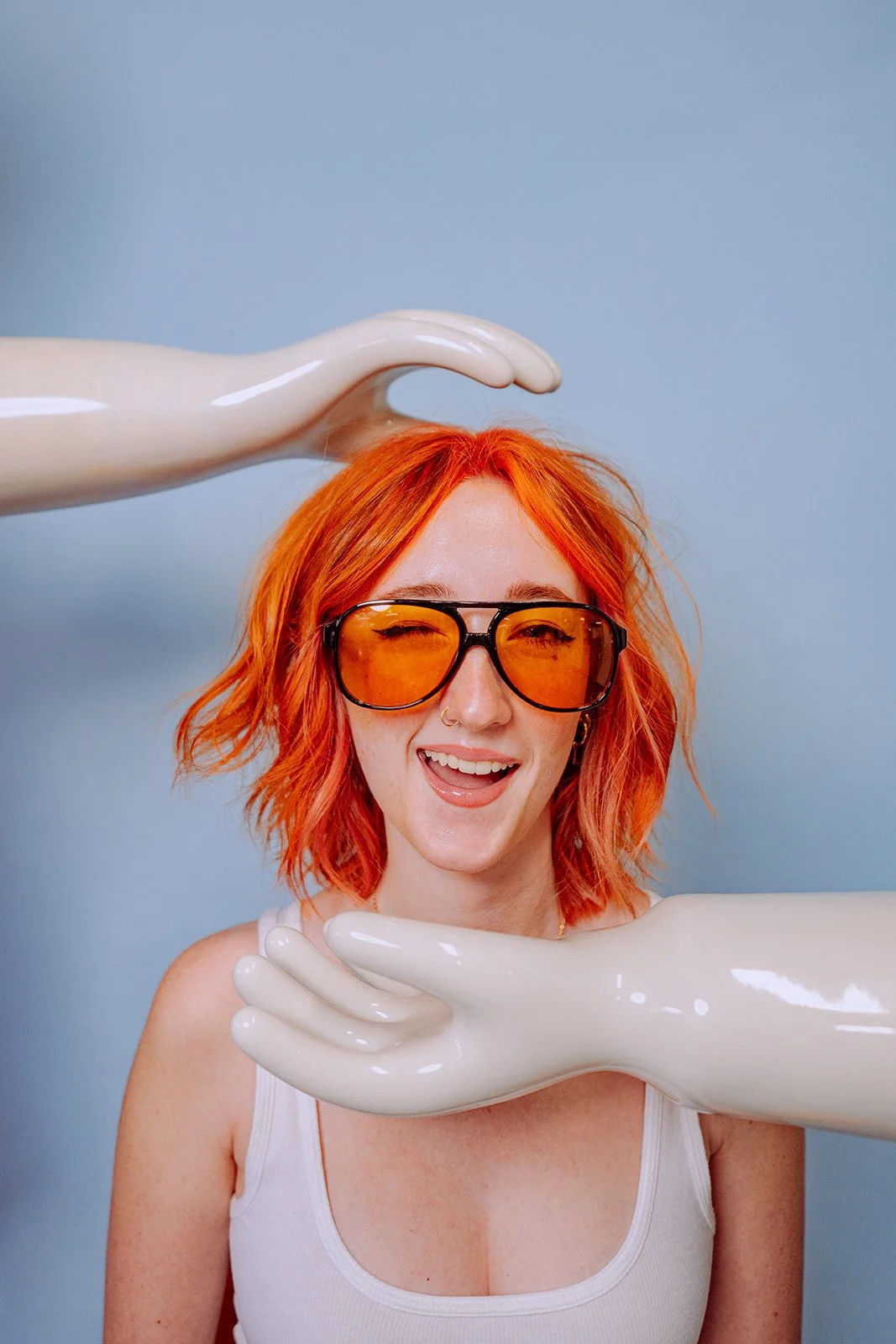 A woman with orange hair wearing yellow-tinted glasses, smiling with her tongue out, is surrounded by two large, white mannequin arms against a blue background.