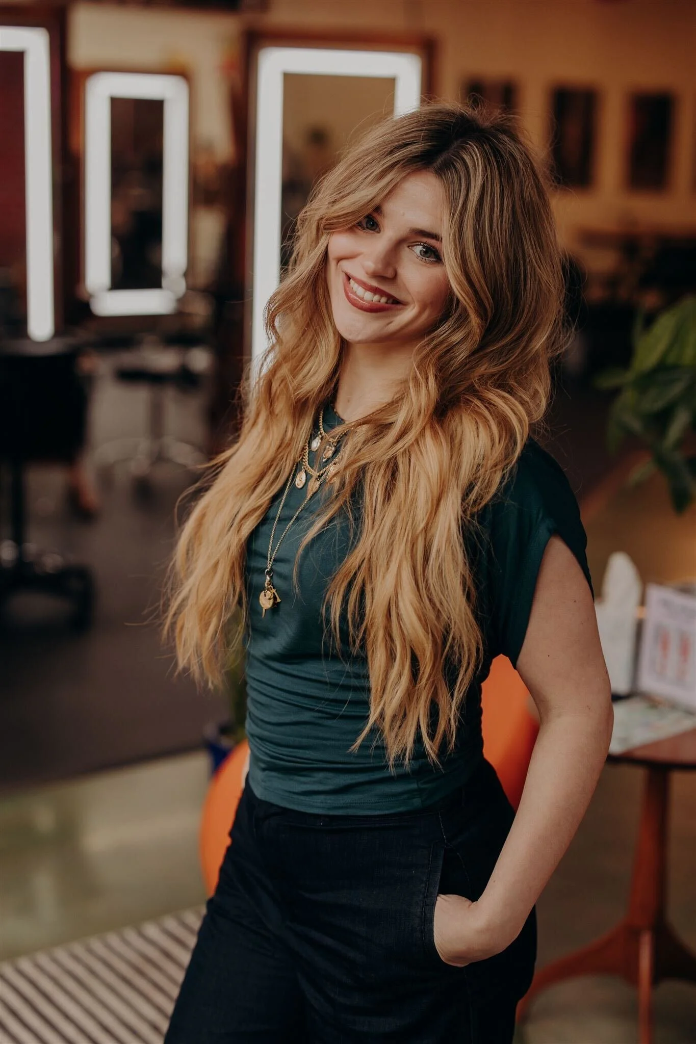 A young woman with long, wavy blonde hair smiling at the camera, wearing a floral ruffled off-the-shoulder top and dark jeans against a blue background.