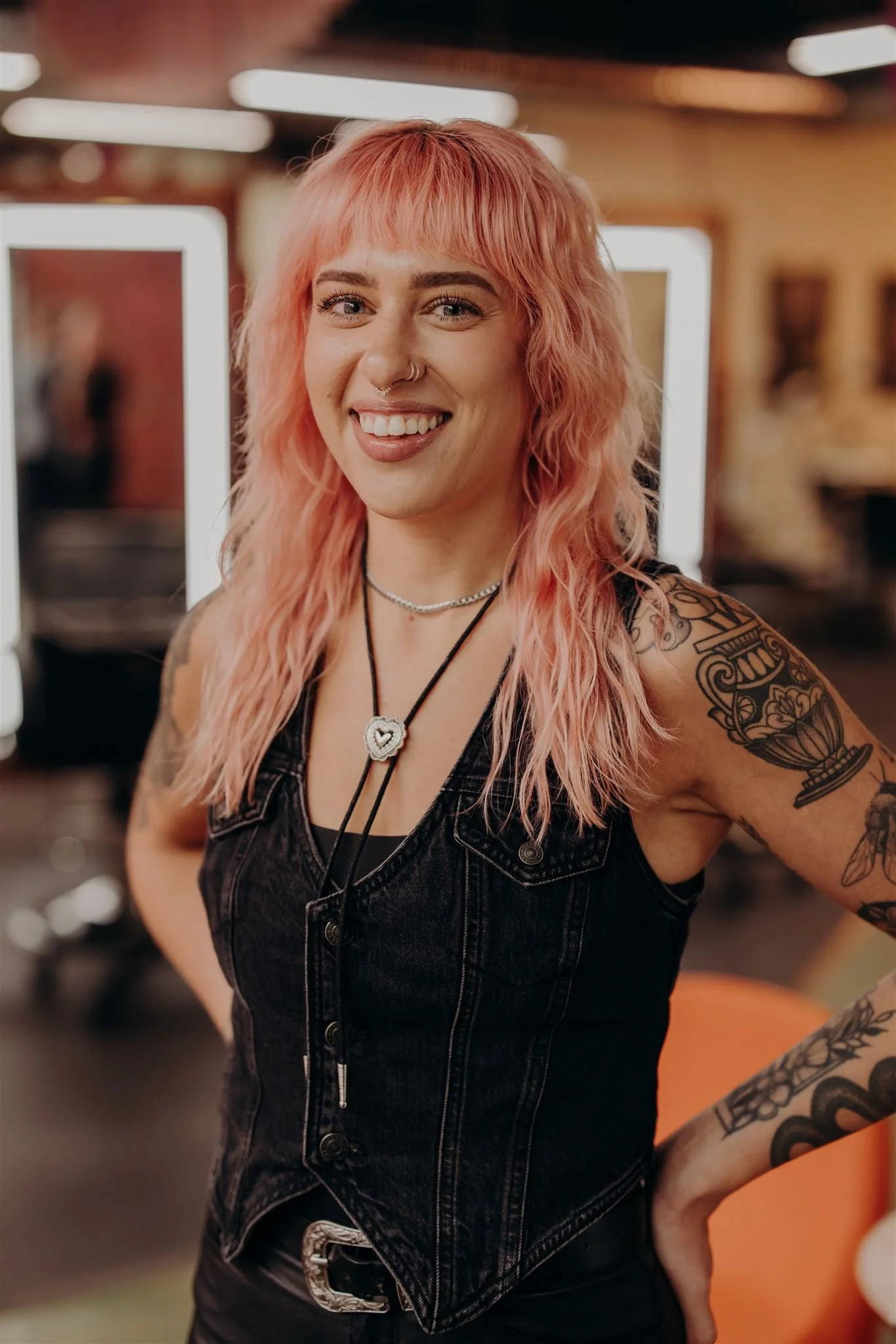 A smiling woman with pink and platinum blonde hair outdoors, wearing a leopard print top, jewelry, and displaying tattoos on her arms.