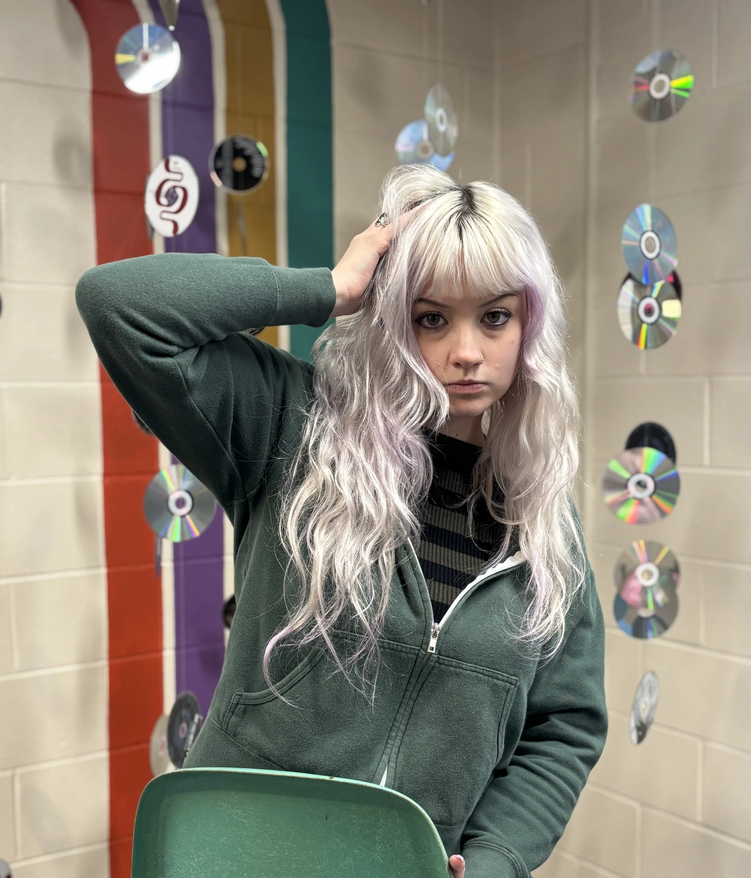 A young woman with long, wavy platinum blonde hair with pastel purple highlights, wearing a green zip-up hoodie over a striped shirt, standing in front of a wall decorated with hanging CDs and a multicolored stripe background, holding a green chair.